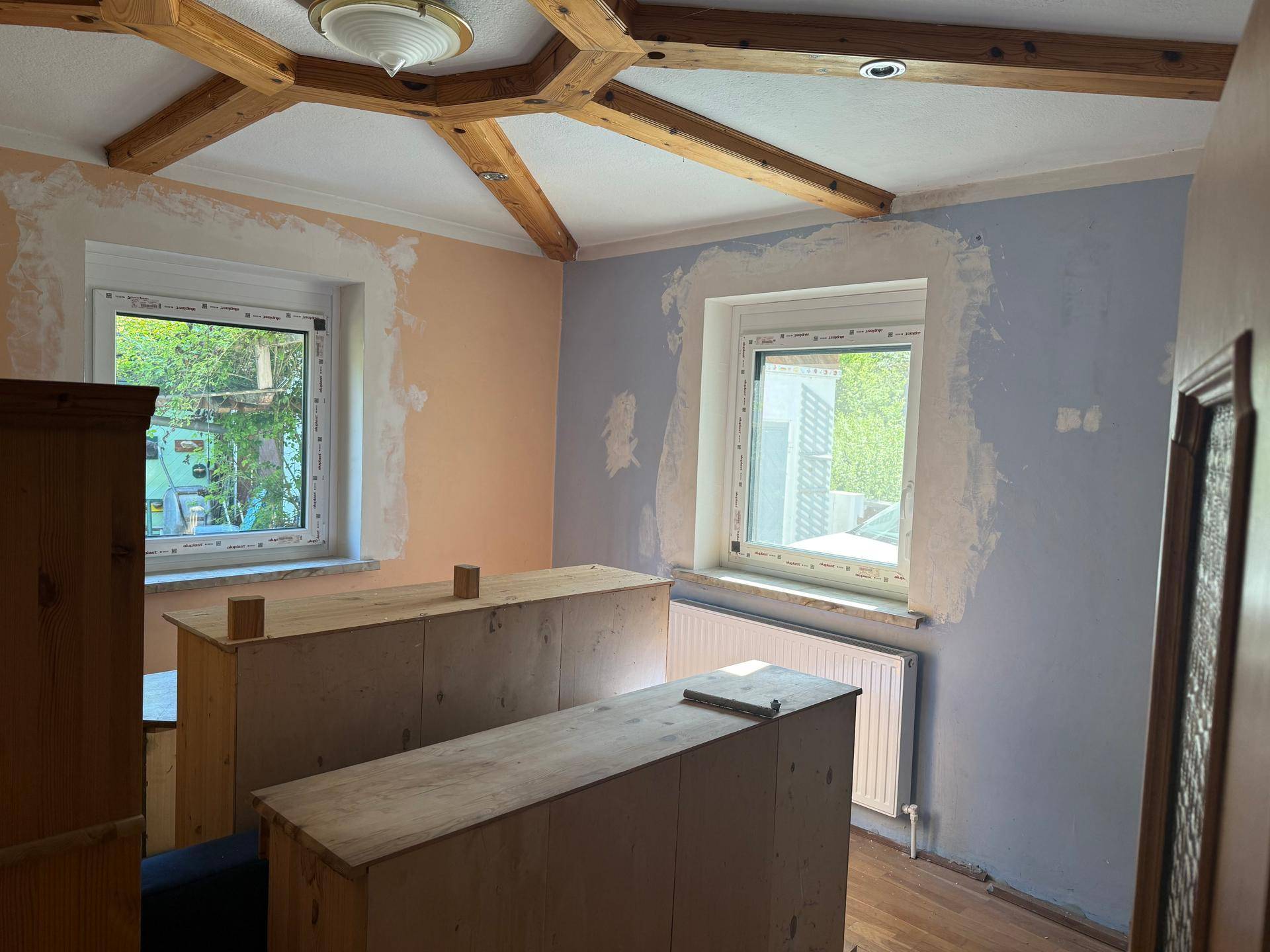 Renovierungsbedürftiger Raum mit sichtbaren Holzbalken an der Decke und zwei Fenstern.