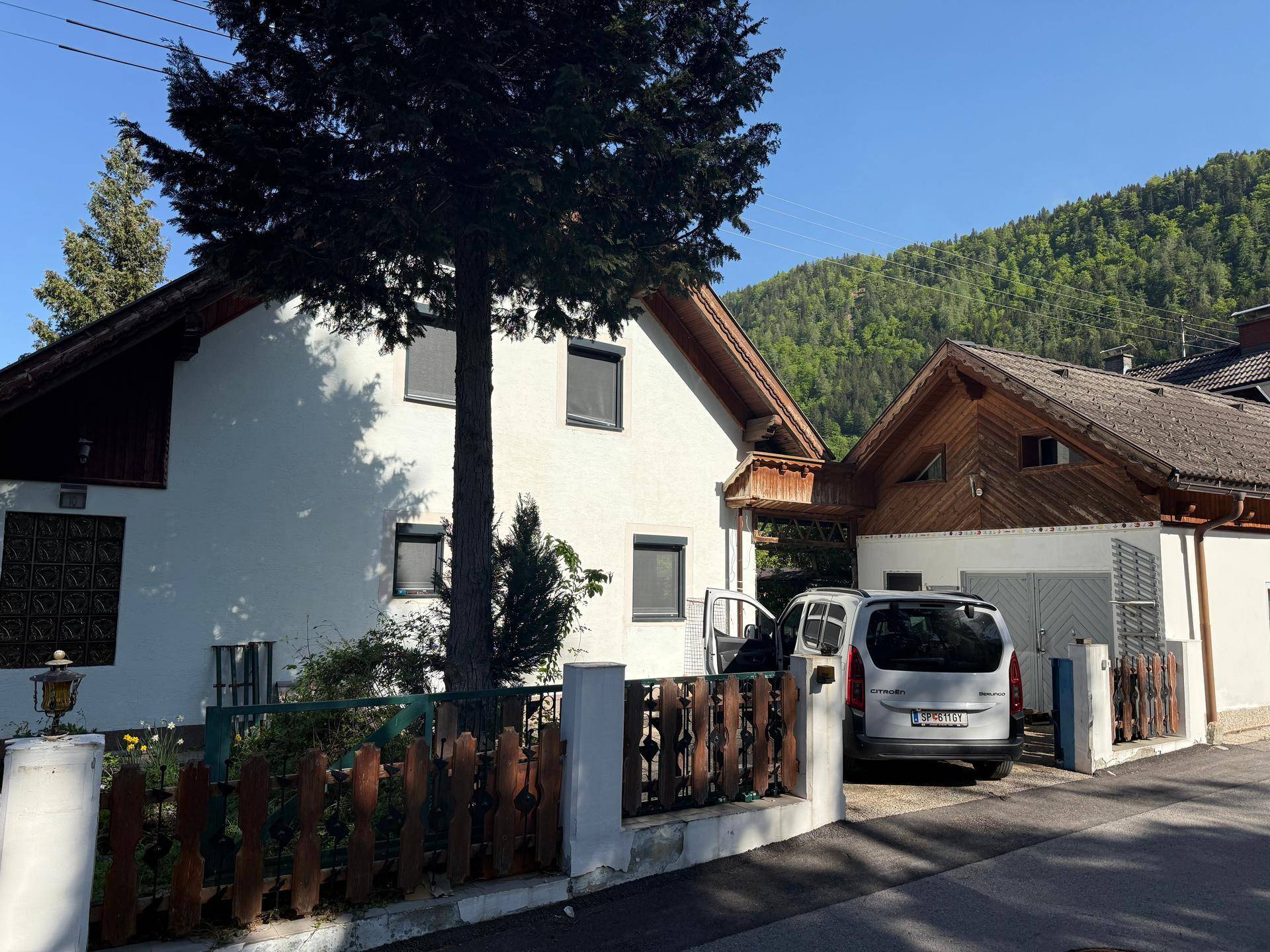 Weißes Einfamilienhaus mit angrenzendem Nebengebäude und Bergblick im Hintergrund.