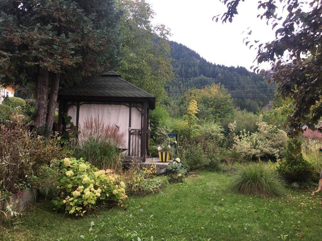 Üppiger Garten mit einem Pavillon, vielfältiger Bepflanzung und bewaldeten Bergen im Hintergrund.