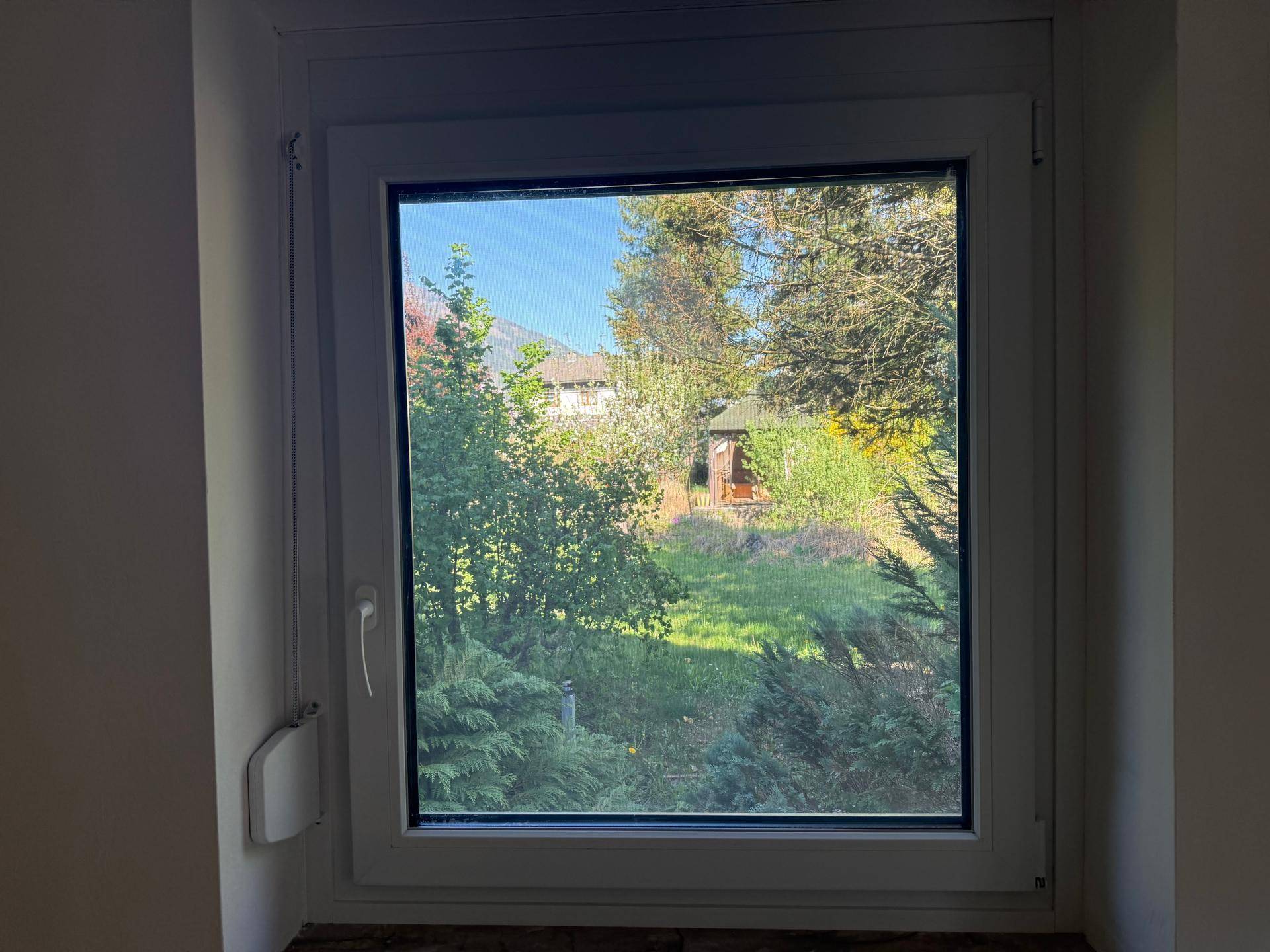 Fensterblick auf einen gepflegten Garten mit Laube und entfernten Bergen.