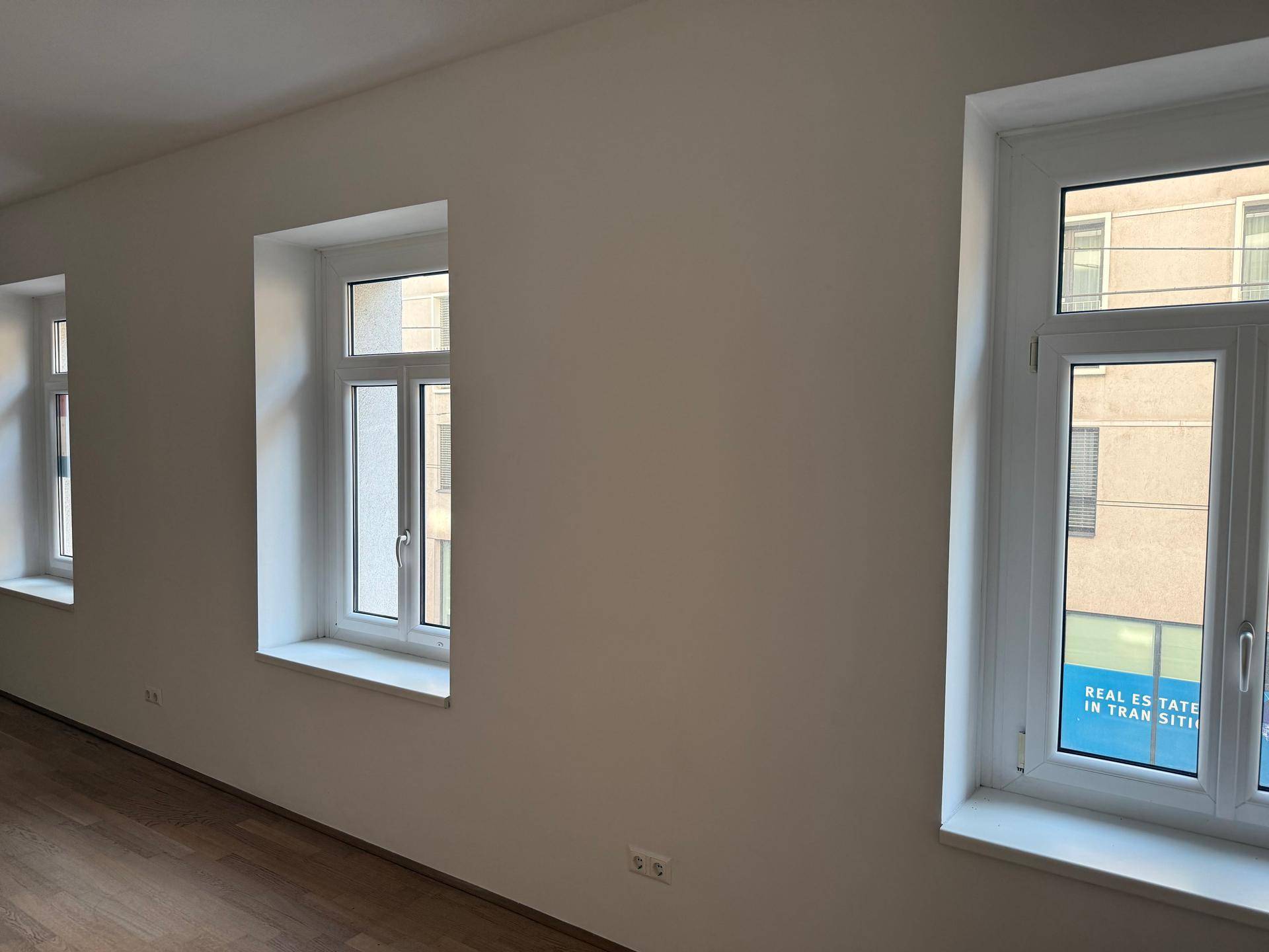 Helle Fensterfront in einem Raum mit Holzfußboden und Blick auf umliegende Gebäude.