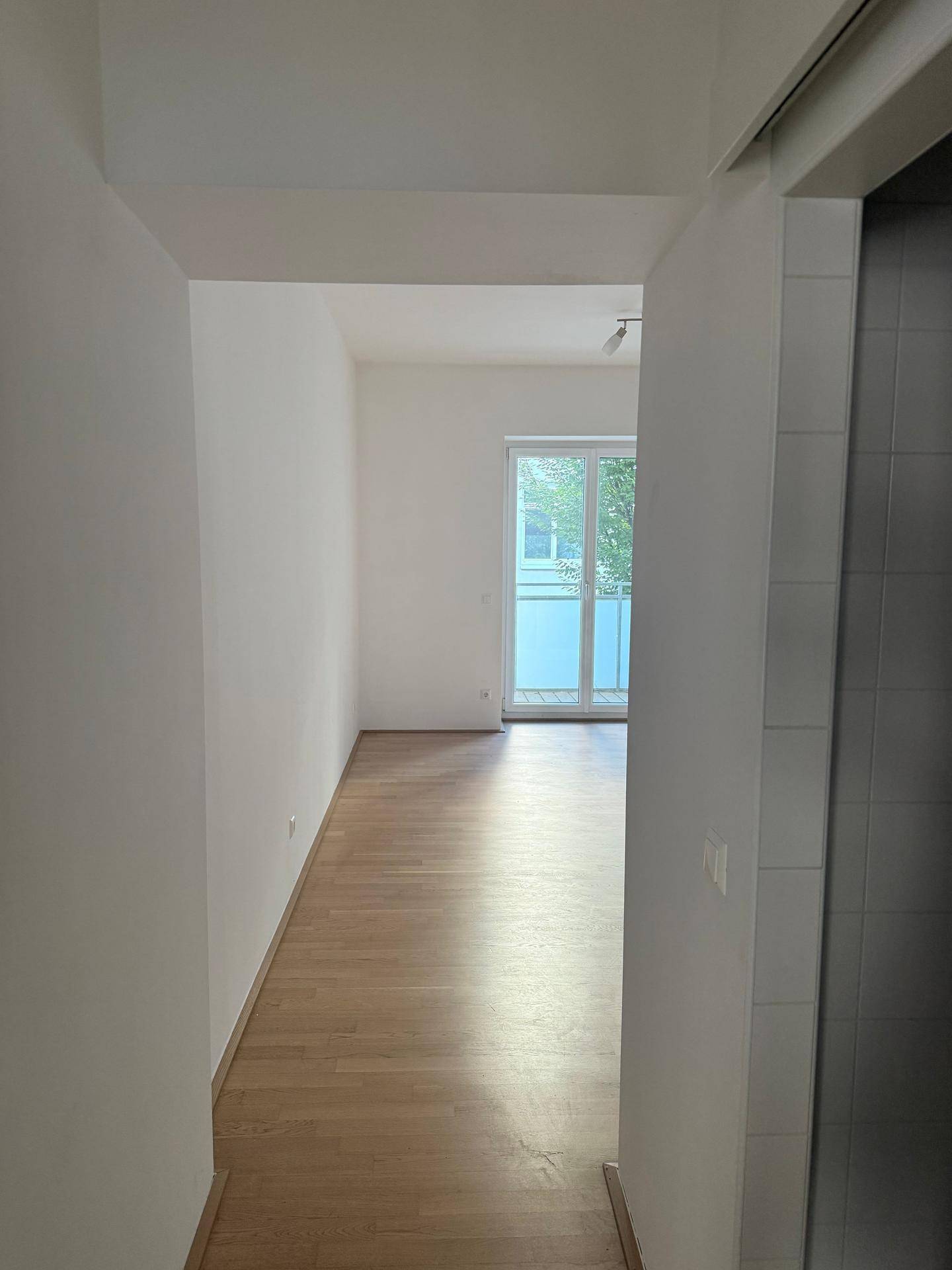 Langer, heller Flur mit Holzfußboden, der zu einem Zimmer mit Balkonzugang führt.