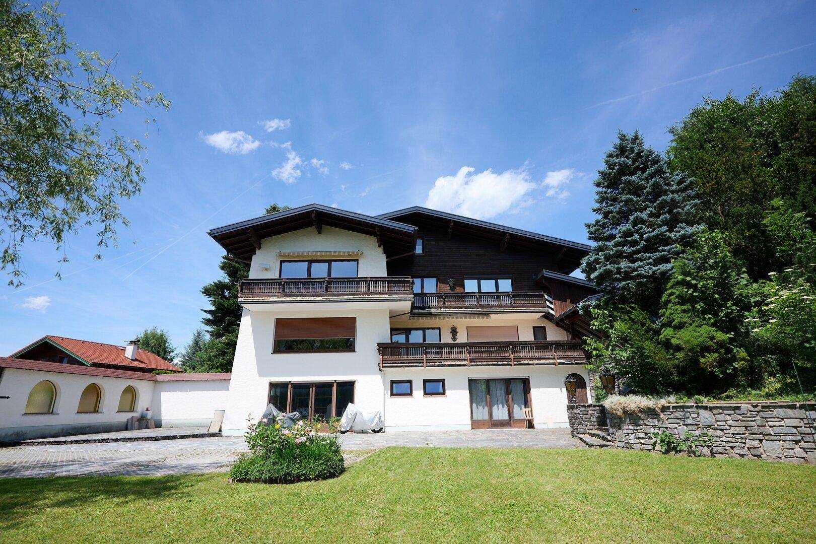 Ansicht des Hauses mit weißer Fassade, Holzbalkonen und gepflegtem Vorgarten unter strahlend blauem Himmel.