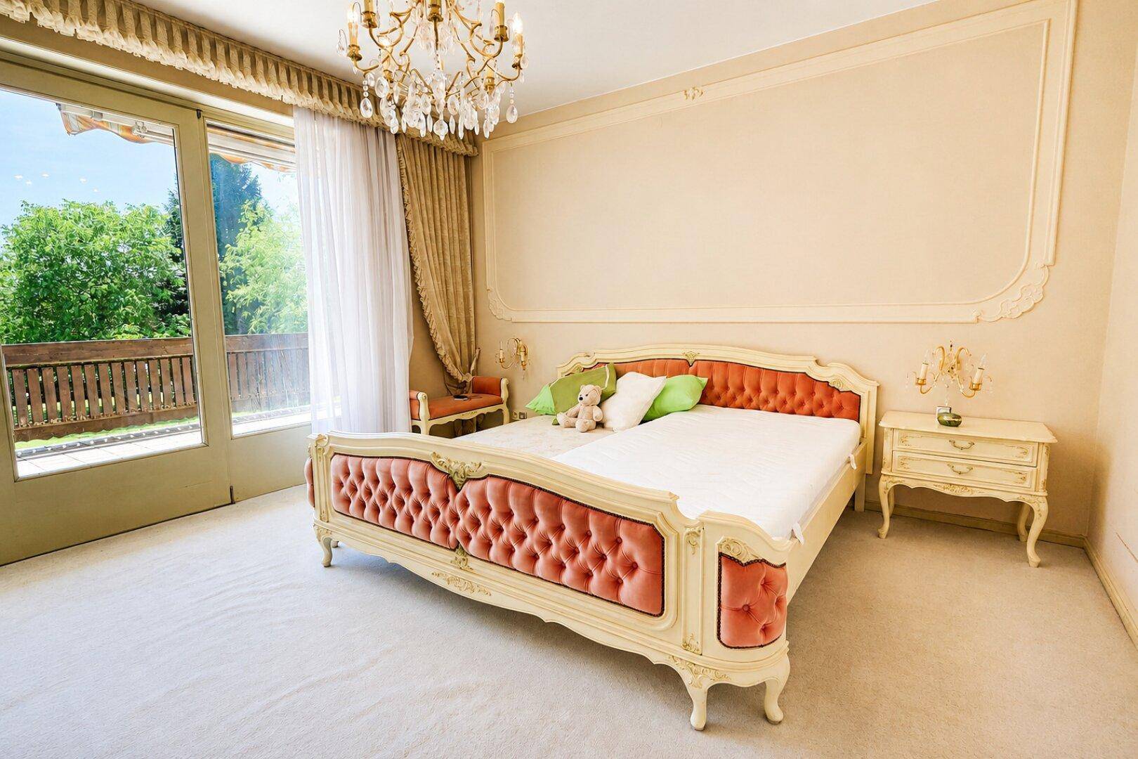 Helles Schlafzimmer mit opulentem Bett, Kronleuchter und Zugang zu einem Balkon mit Blick ins Grüne.