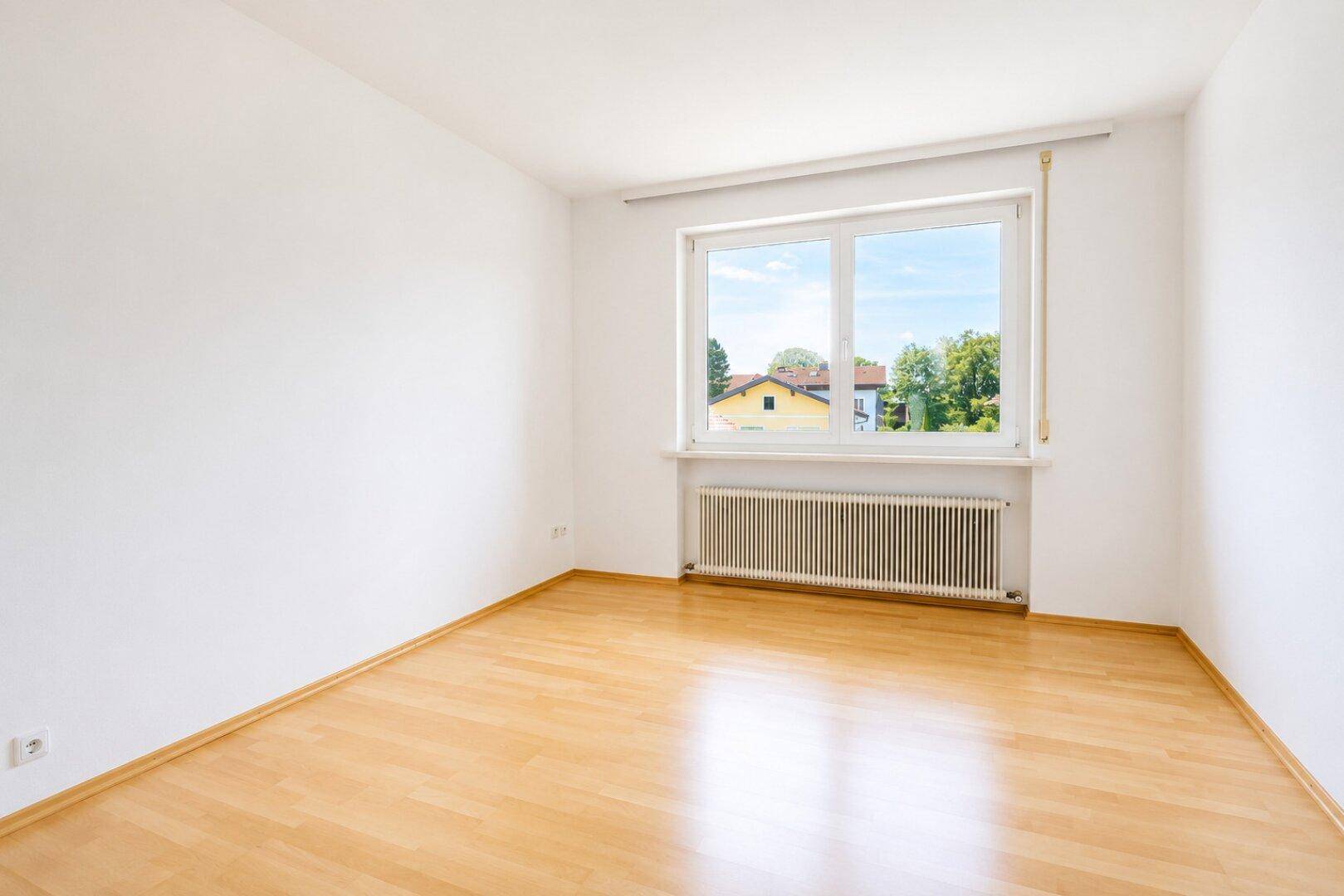 Helles, leeres Schlafzimmer mit Laminatboden und großem Fenster, das viel Tageslicht hereinlässt.