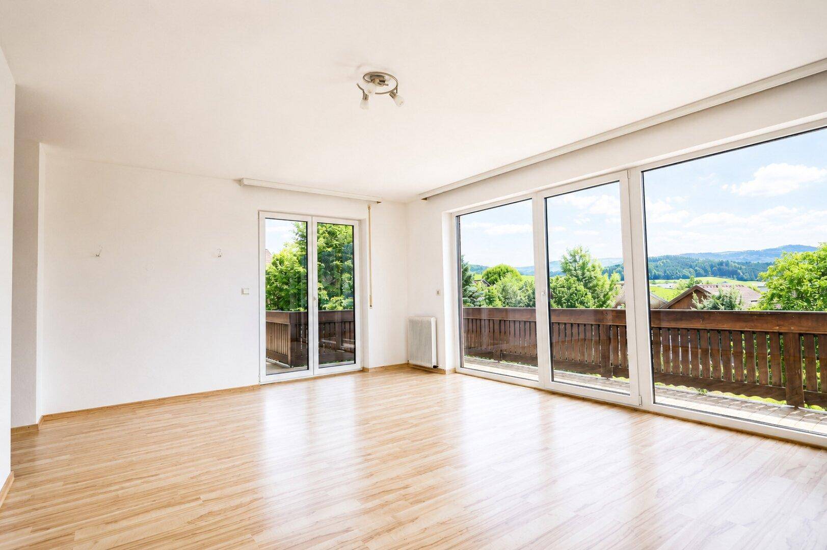 Geräumiges, leeres Zimmer mit großen Fenstern und Balkonzugang, bietet einen weiten Blick in die Landschaft.