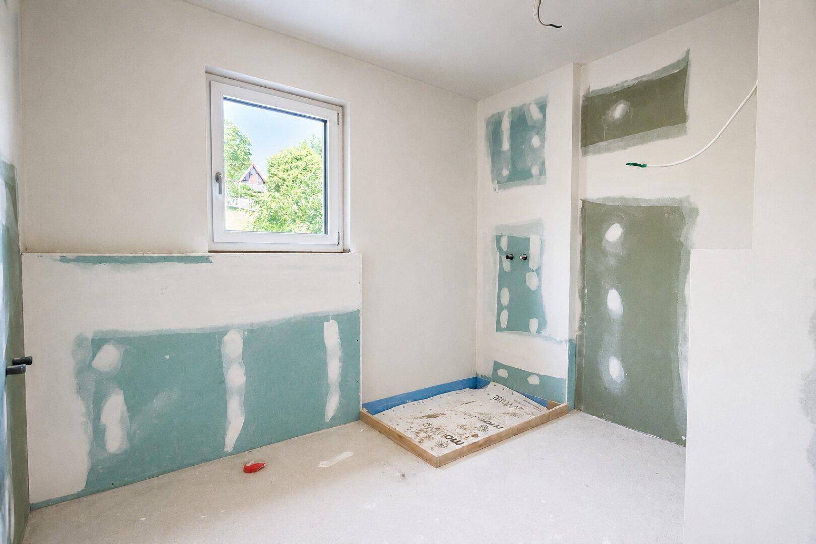 Badezimmer im Rohbau mit vorbereiteter Duschfläche und Fenster für natürliche Belüftung.
