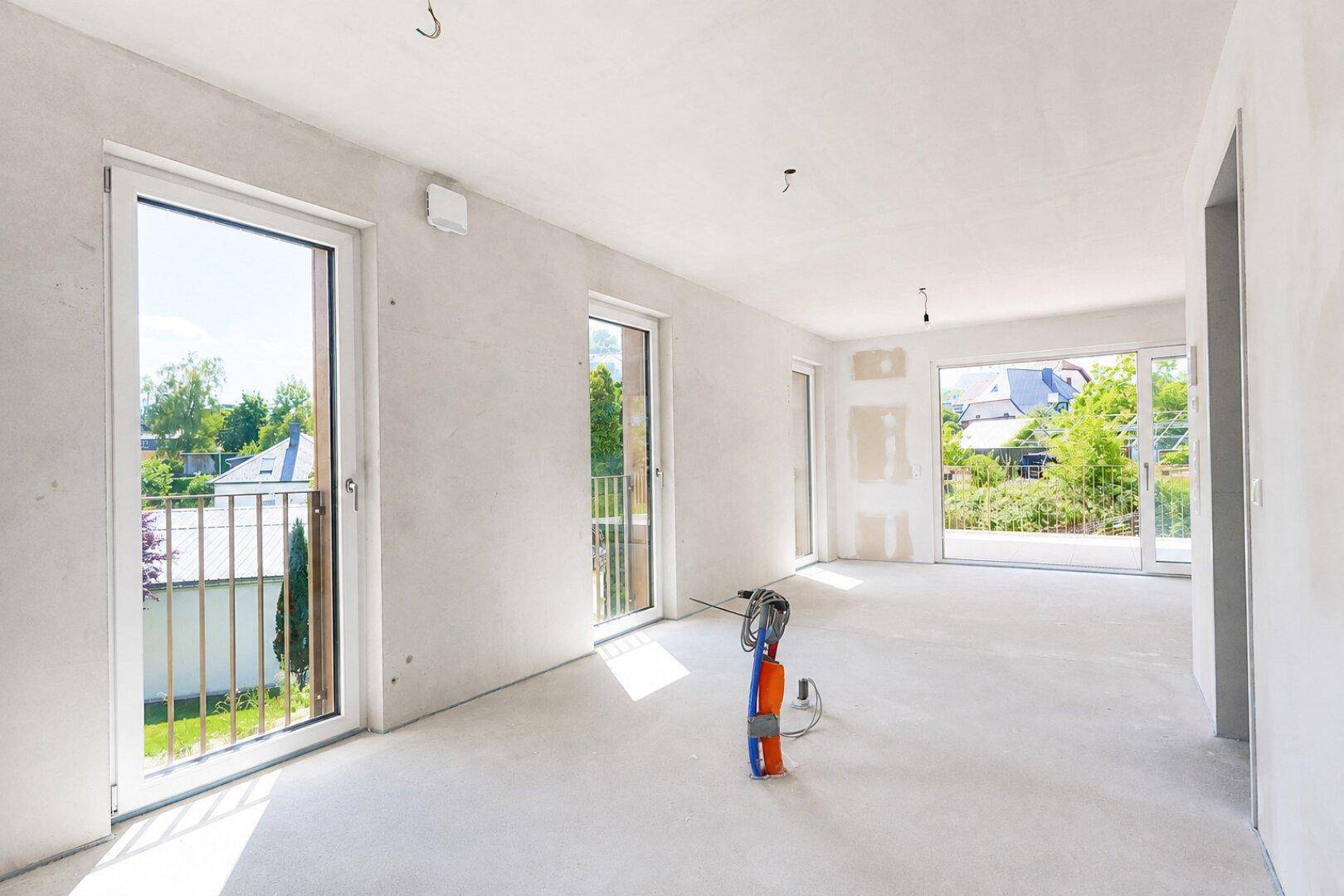 Geräumiger Wohnbereich im Rohbau mit großen Fenstern und Zugang zu einem französischen Balkon.