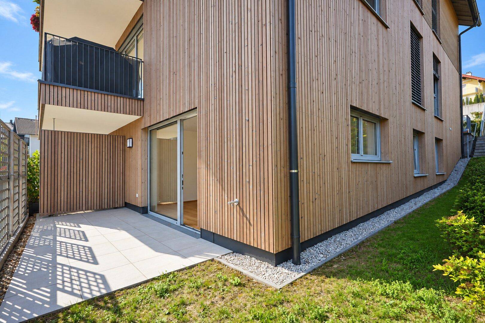 Moderne Holzfassade eines Neubaus mit sonniger Terrasse, kleinem Garten und Balkon im Obergeschoss.