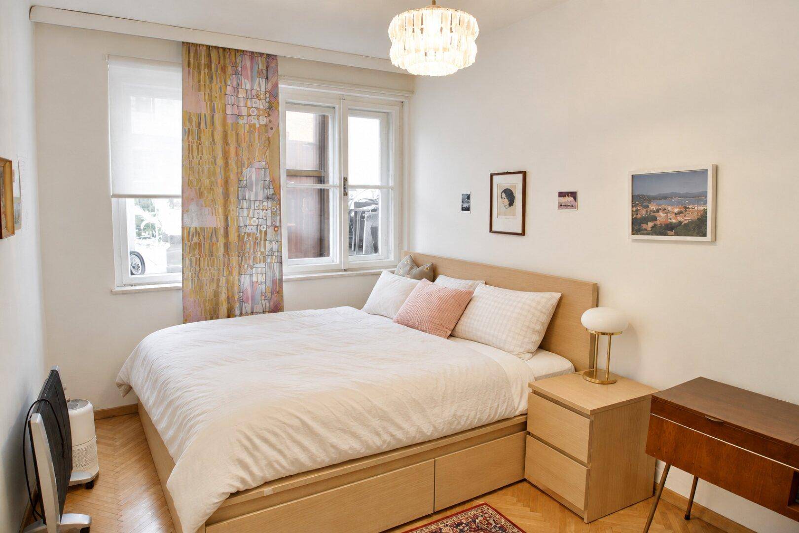Gemütliches Schlafzimmer mit Doppelbett, Holzboden und einem modernen Kronleuchter, hell beleuchtet.