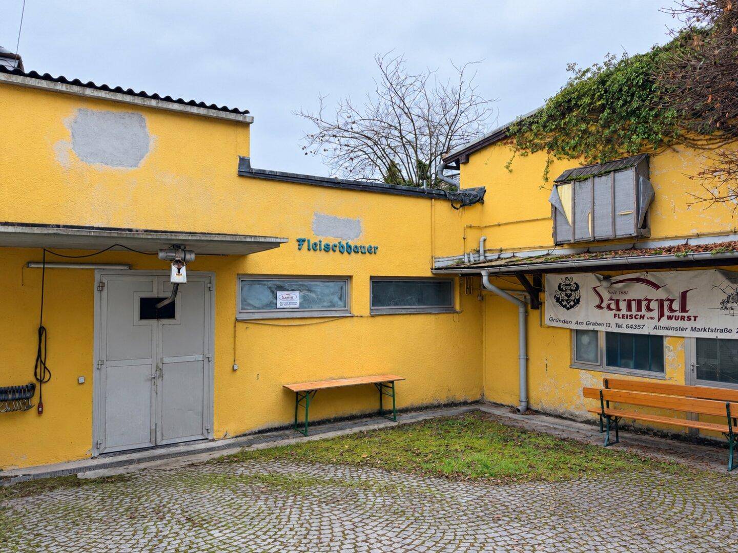 Gelbes Gebäude mit einem gepflasterten Hinterhofbereich, Bänken und Hinweisschildern.