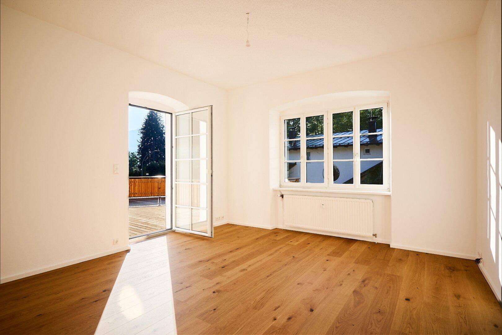 Heller Raum mit Holzboden und Zugang zu einer Terrasse, mit Blick ins Grüne.