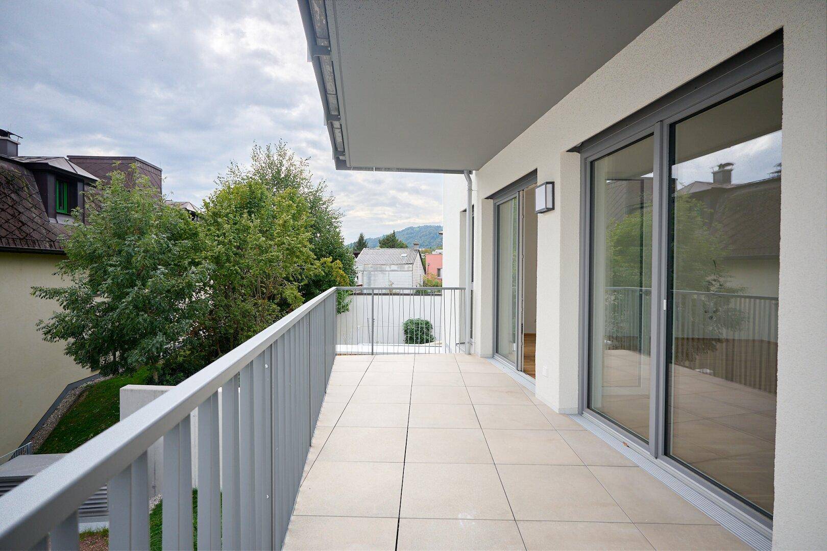 Langer Balkon mit Fliesenboden und Blick auf Bäume und die Umgebung.