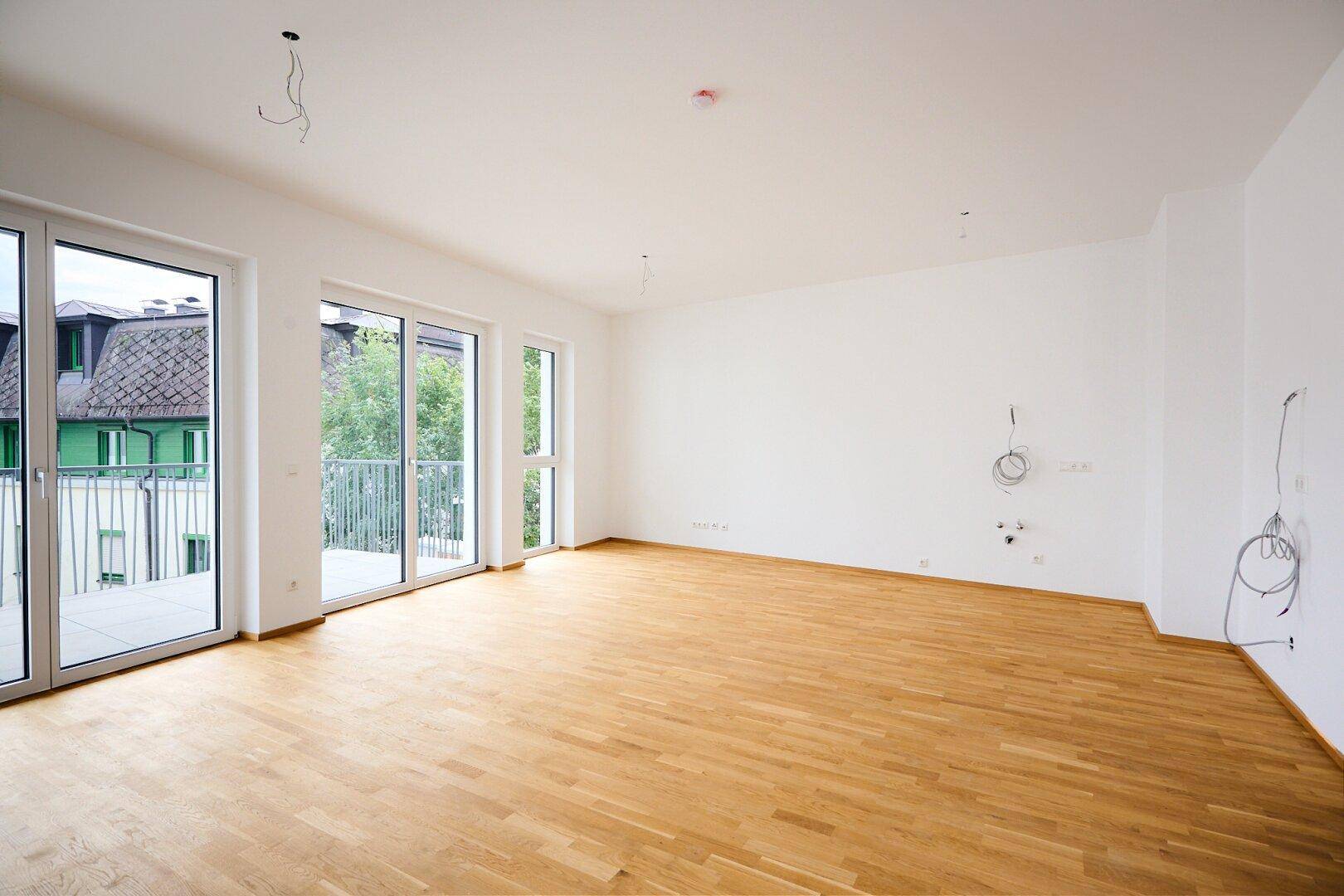 Geräumiges, leeres Wohnzimmer mit Parkettboden und Zugang zu einem Balkon.