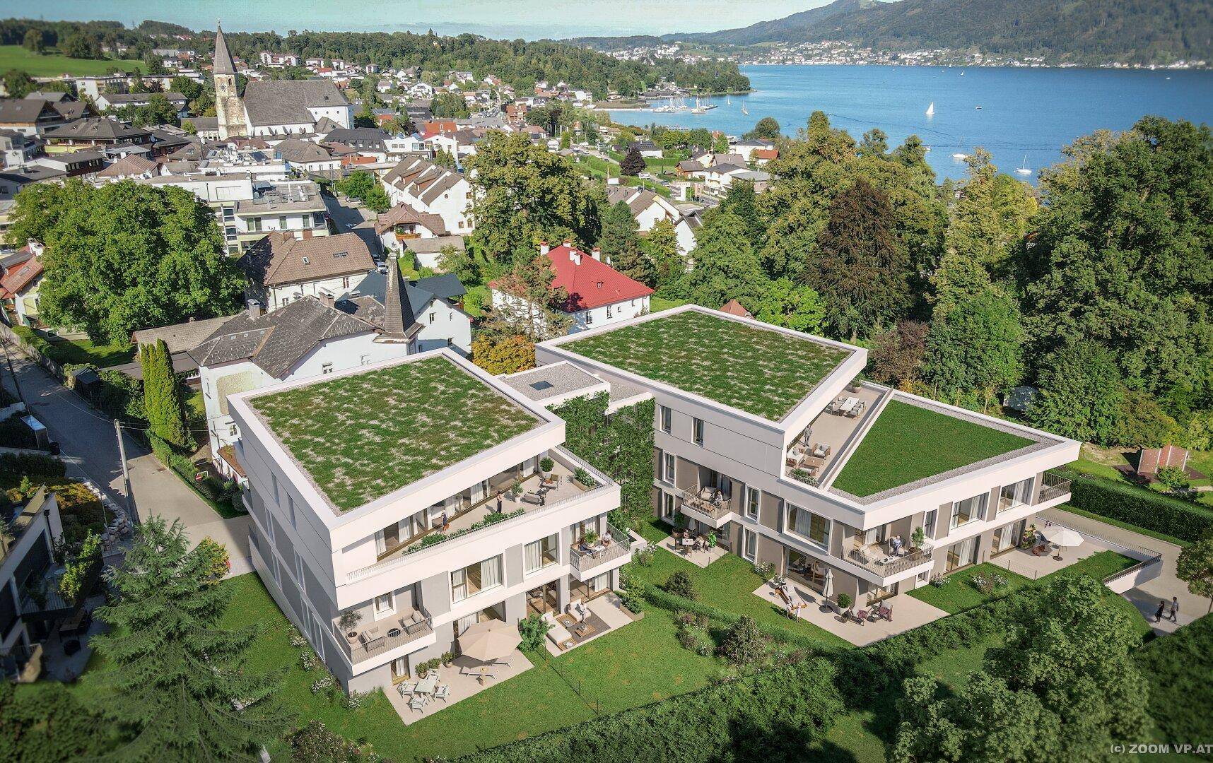 Luftaufnahme der modernen Wohnanlage mit See, Bergen und historischer Kirche im Hintergrund.