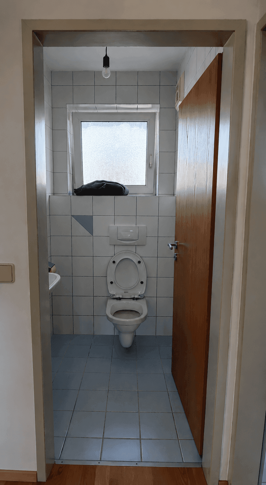 Separates WC mit weißen Fliesen, blauem Boden, wandhängender Toilette und einem kleinen Fenster.