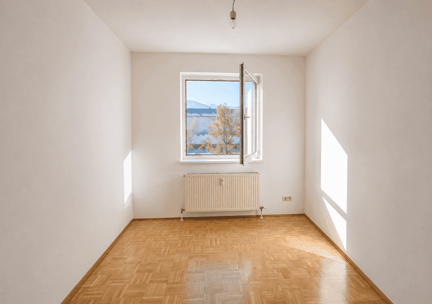 Helles, leeres Schlafzimmer mit Parkettboden, weißen Wänden und einem Fenster mit Heizkörper.