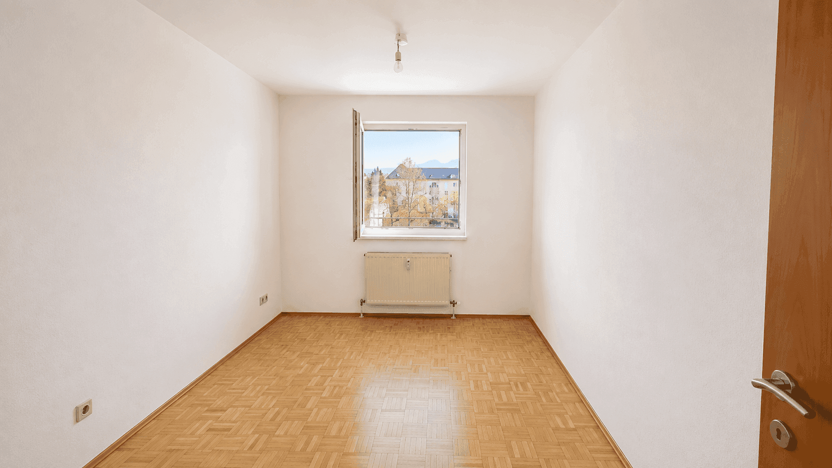 Leeres Schlafzimmer mit hellem Parkettboden, weißen Wänden und einem Fenster, das Tageslicht hereinlässt.