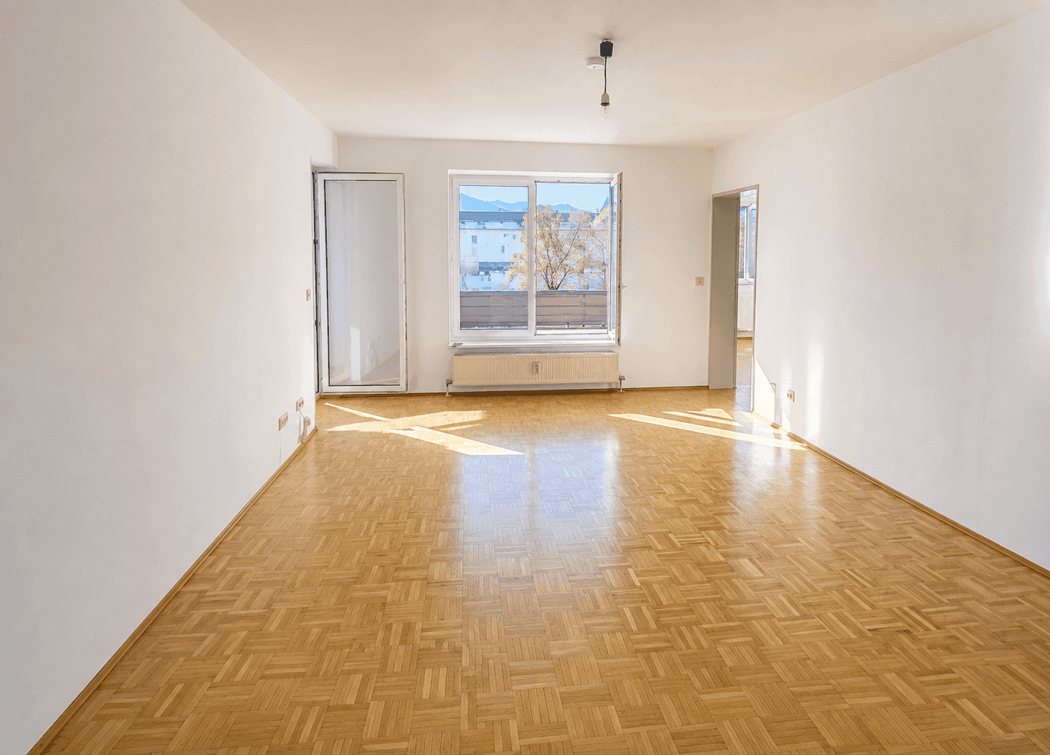 Helles, leeres Wohnzimmer mit Fischgrätparkettboden, weißen Wänden und Zugang zu einem Balkon.