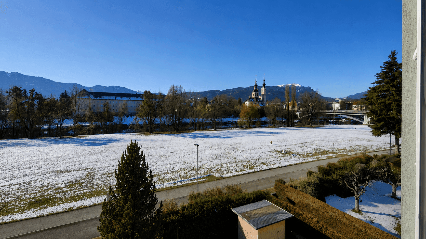 Winterlandschaft mit schneebedecktem Feld, Bergen im Hintergrund und einem historischen Gebäude unter blauem Himmel.