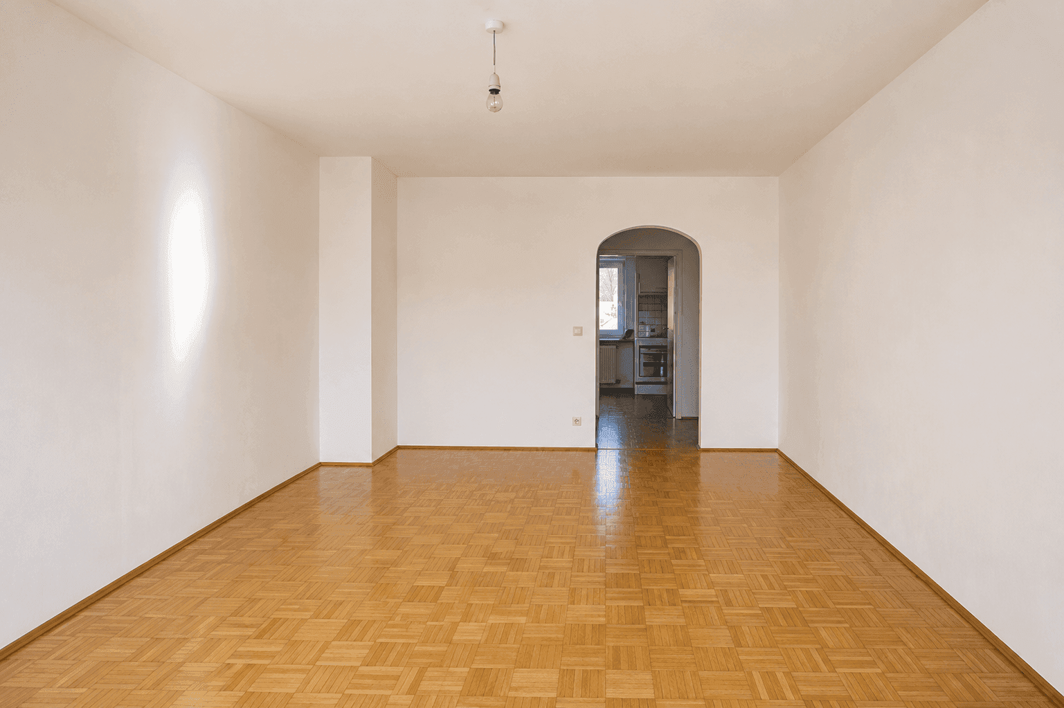 Geräumiges, leeres Wohnzimmer mit Parkettboden und einem Rundbogen, der zur Küche führt.