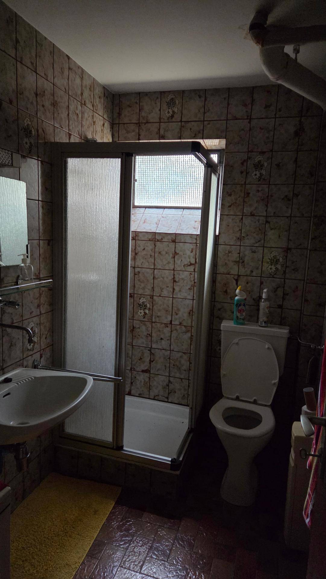 Kleines Badezimmer mit dunklen Fliesen, Duschkabine, WC und Waschbecken.
