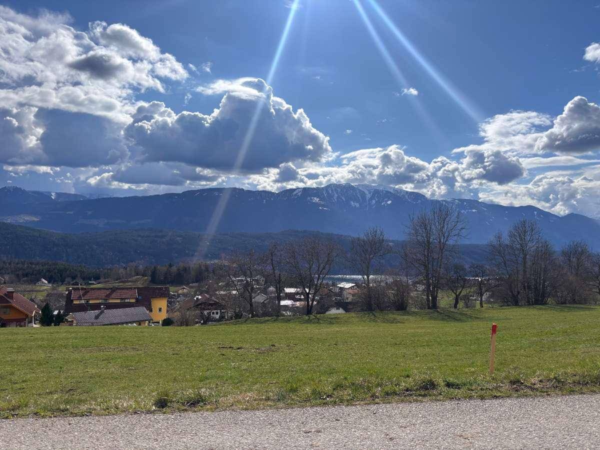 Sonnige Landschaft mit grüner Wiese, Häusern und schneebedeckten Bergen unter blauem Himmel.