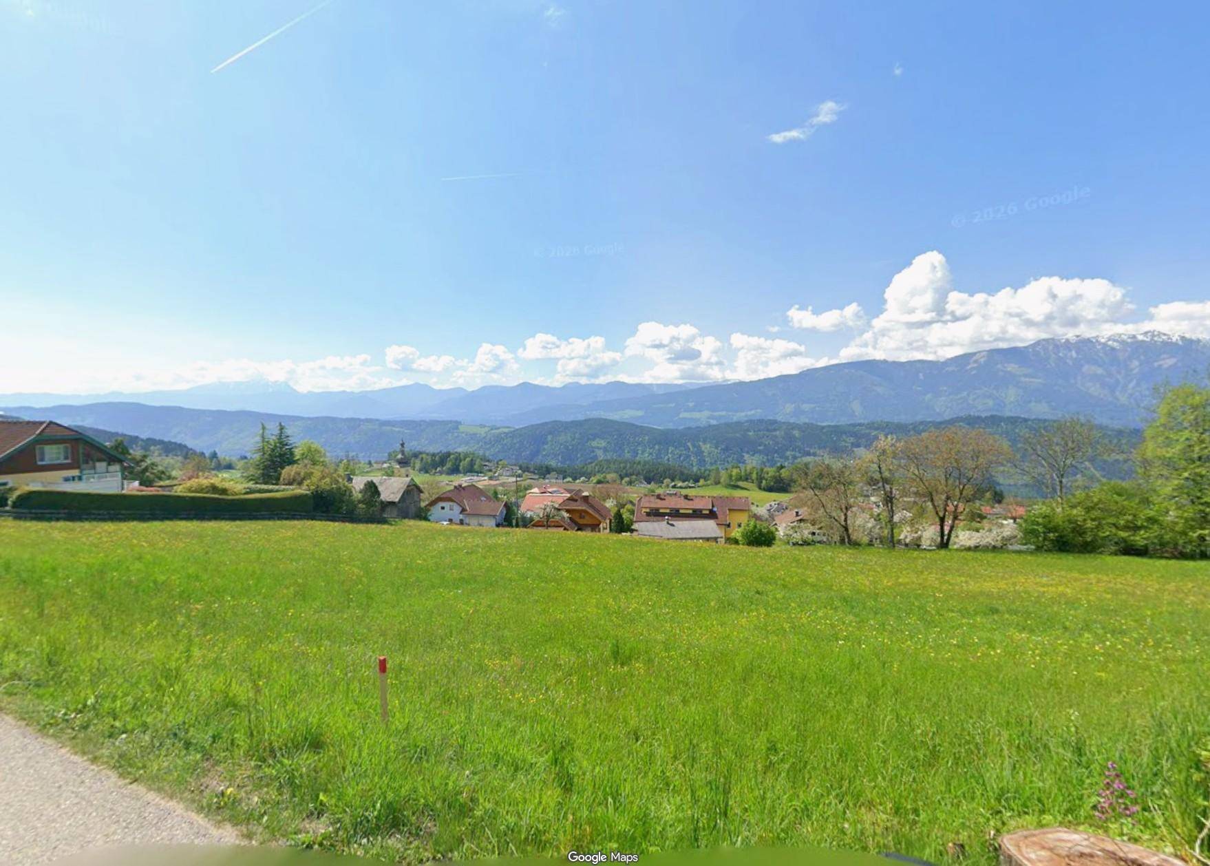 Weitläufige grüne Wiese mit Blick auf eine Wohnsiedlung und die umliegende Berglandschaft.