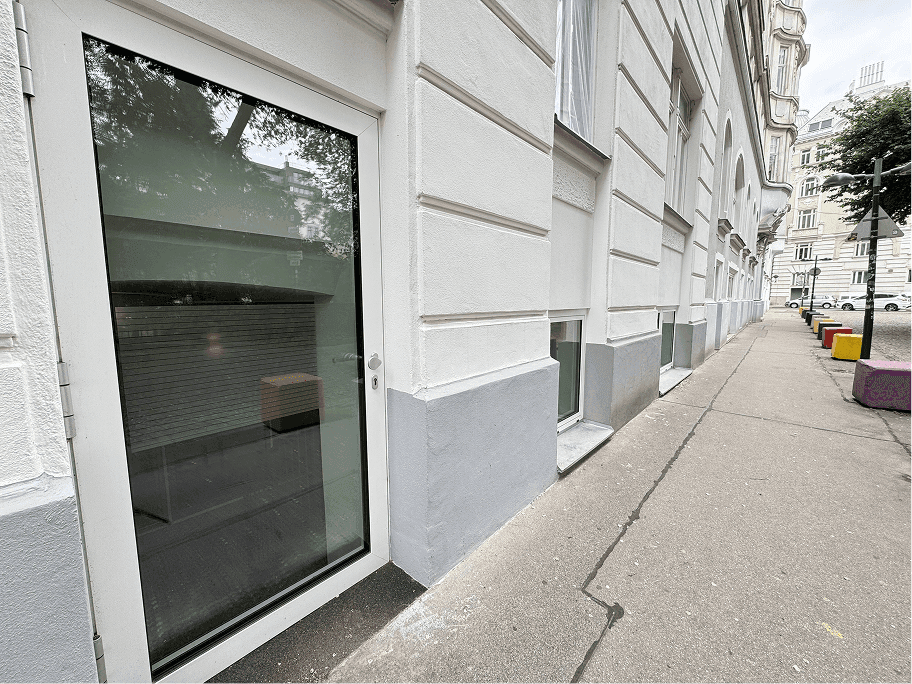 Seitenansicht eines Gebäudes mit großen Fenstern im Erdgeschoss und einem Bürgersteig.