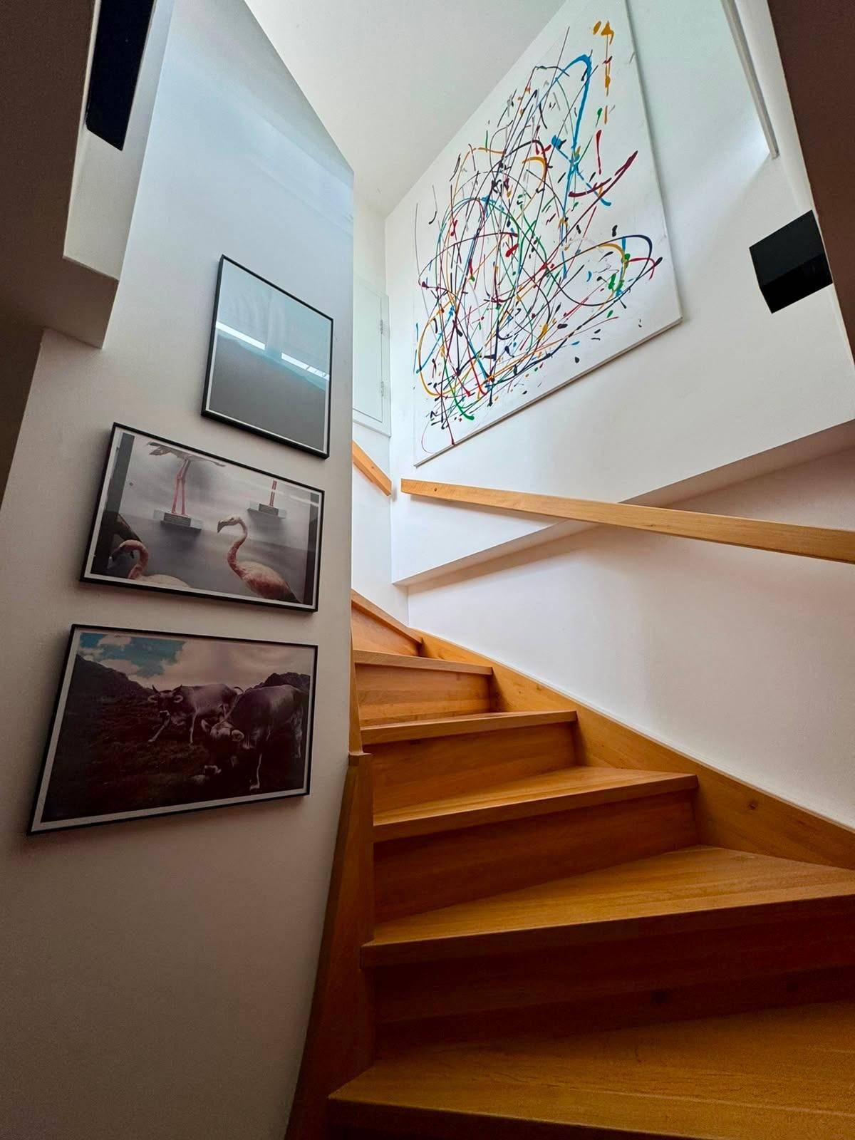 Helle Holztreppe mit Geländer und einer Galerie von modernen Kunstwerken an der Wand.