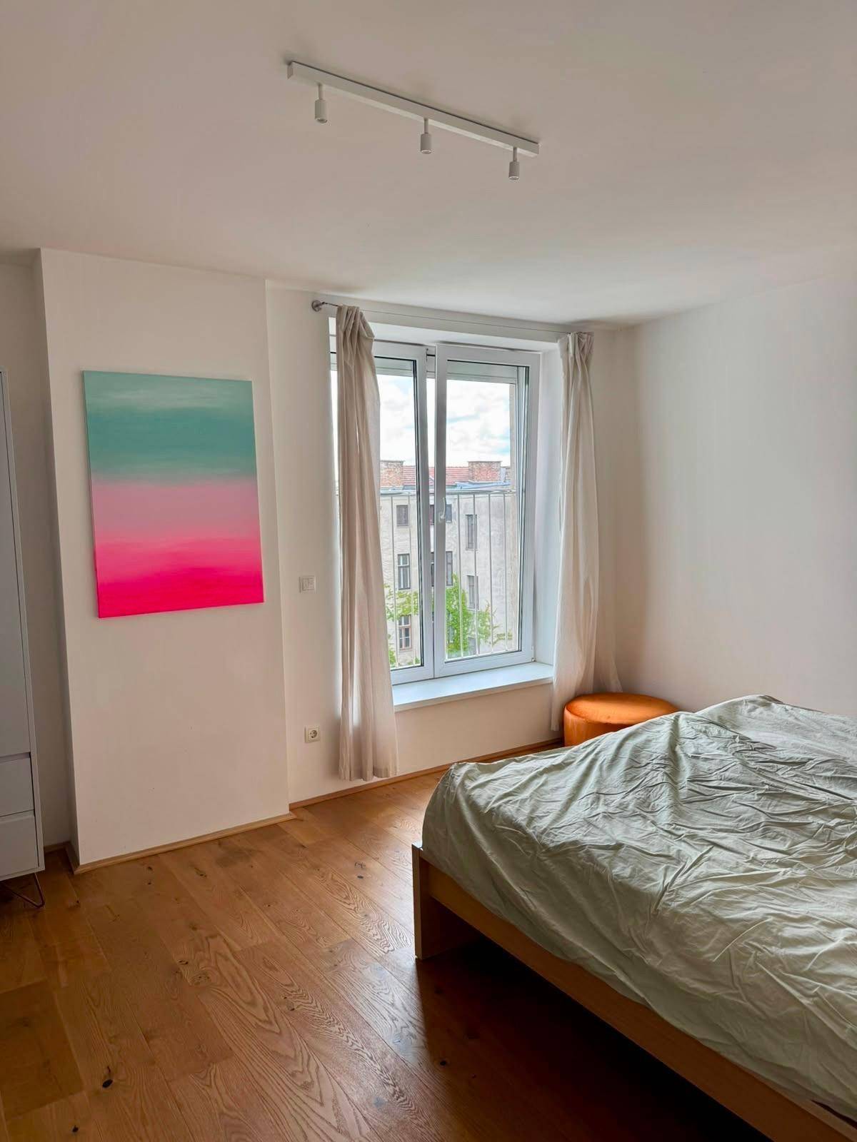 Helles Schlafzimmer mit Holzbett, großem Fenster und einem farbenfrohen Kunstwerk an der Wand.