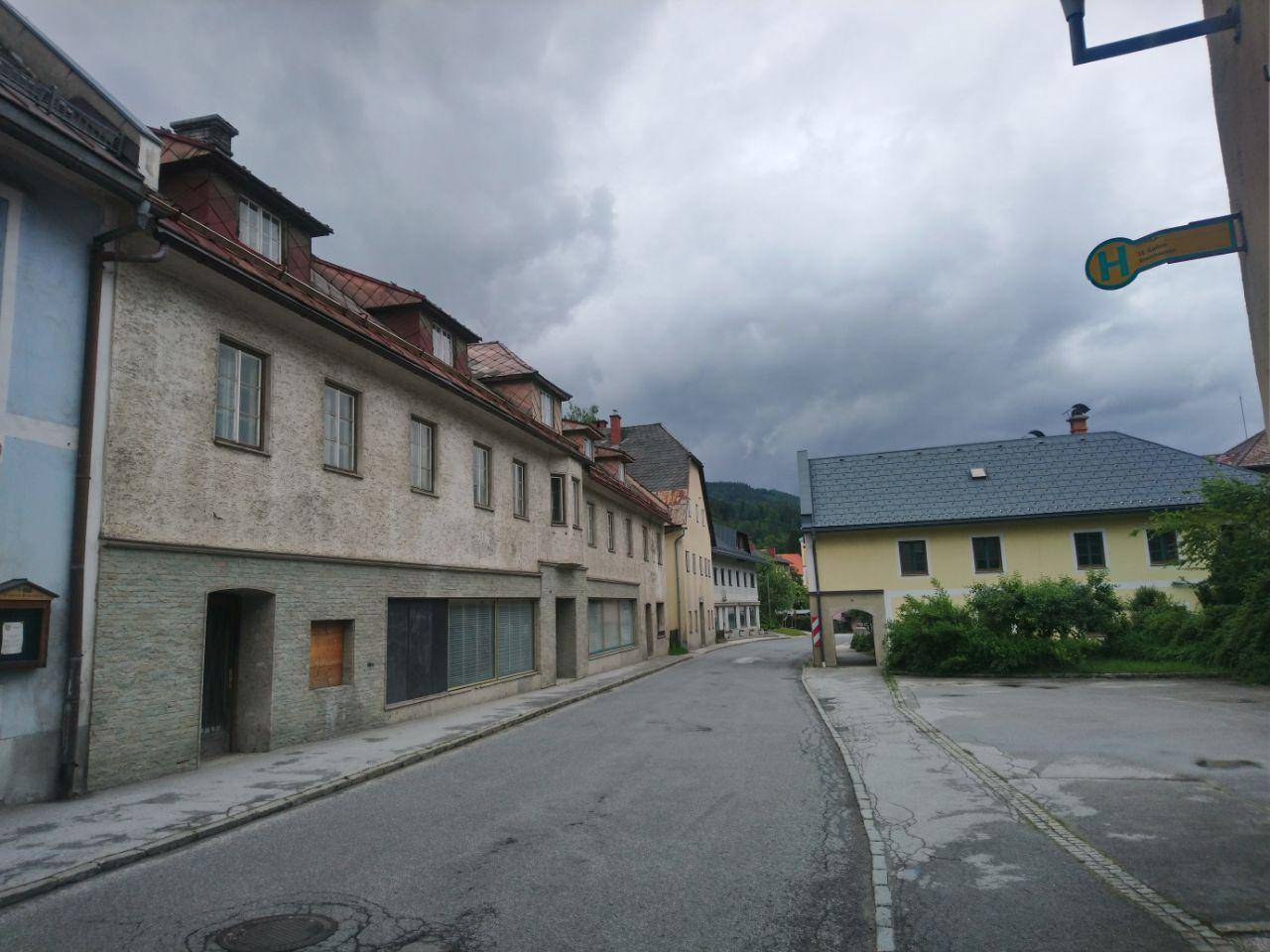 Blick auf eine ruhige Straße mit älteren Gebäuden und bewaldeten Hügeln im Hintergrund unter bewölktem Himmel.