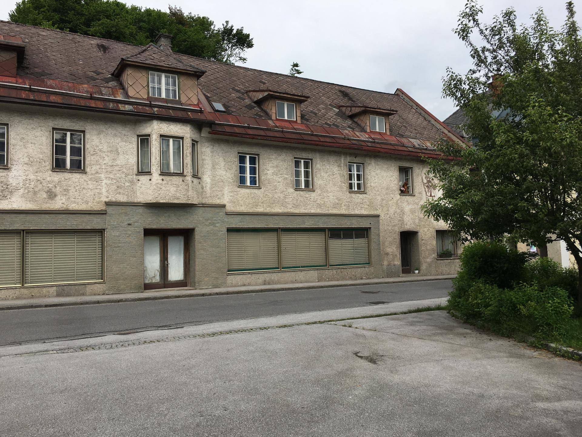 Ansicht eines älteren Gebäudes mit mehreren Fenstern und Ladenfronten an einer Straße, umgeben von Bäumen.