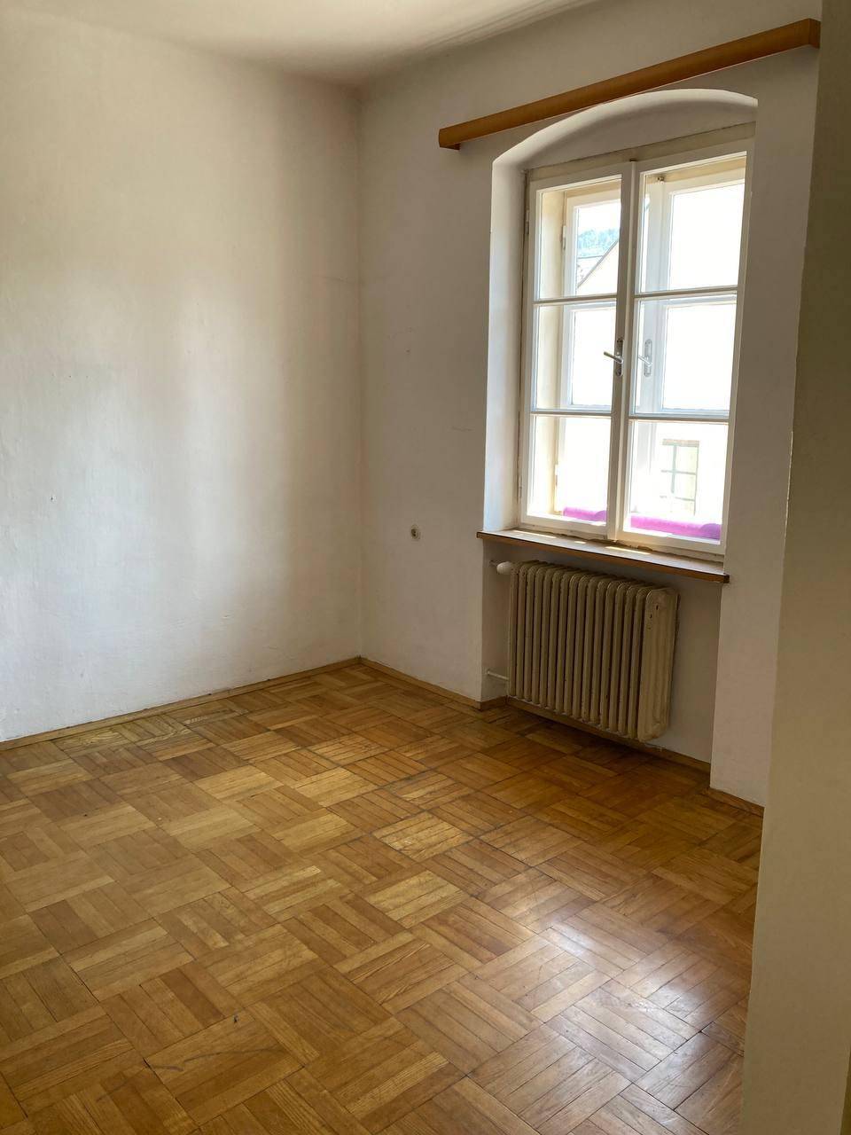Helles, leeres Zimmer mit klassischem Fischgrätparkett, weißen Wänden und einem Fenster mit Heizkörper.