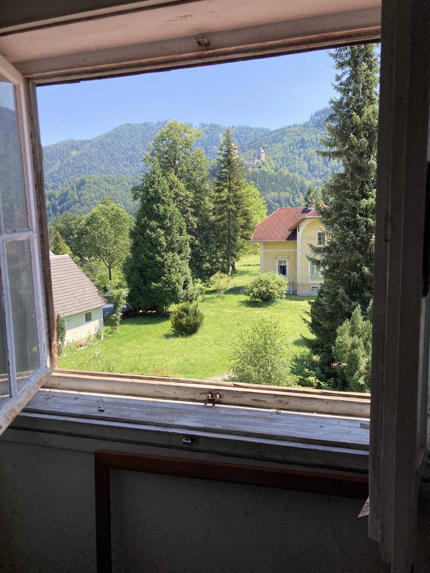 Offenes Fenster bietet einen weiten Blick auf grüne Hügel, Bäume und ein Haus mit rotem Dach in der Ferne.
