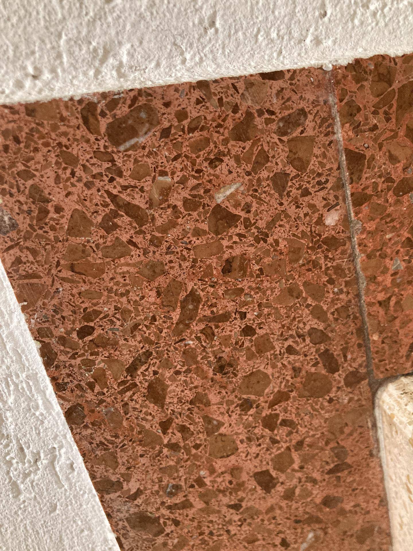 Nahaufnahme eines roten Terrazzobodens mit unregelmäßigen Steinsplittern und weißer Wandstruktur.