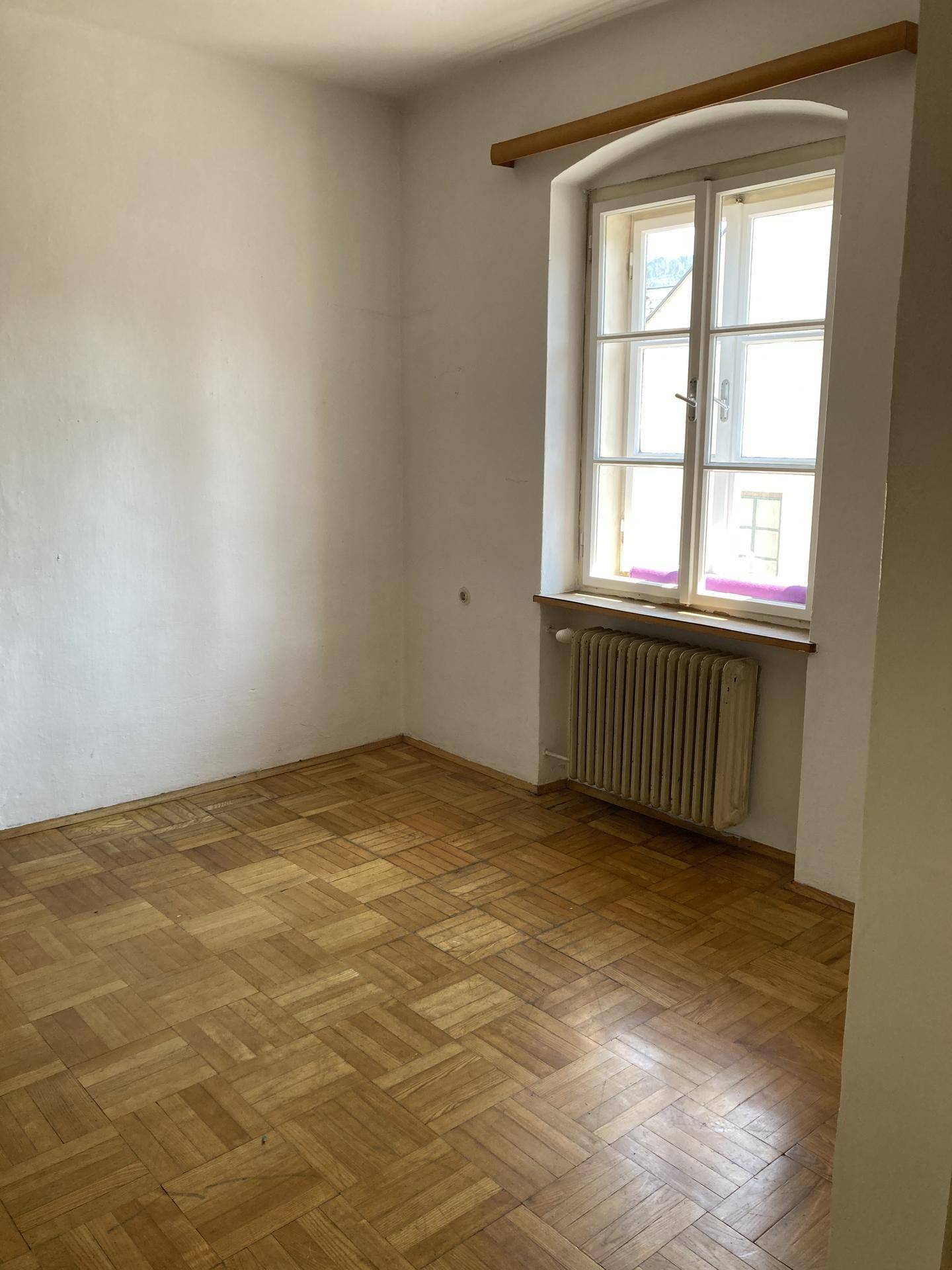Leeres Zimmer mit Fischgrätparkett, weiß gestrichenen Wänden und einem Fenster mit Heizkörper darunter.