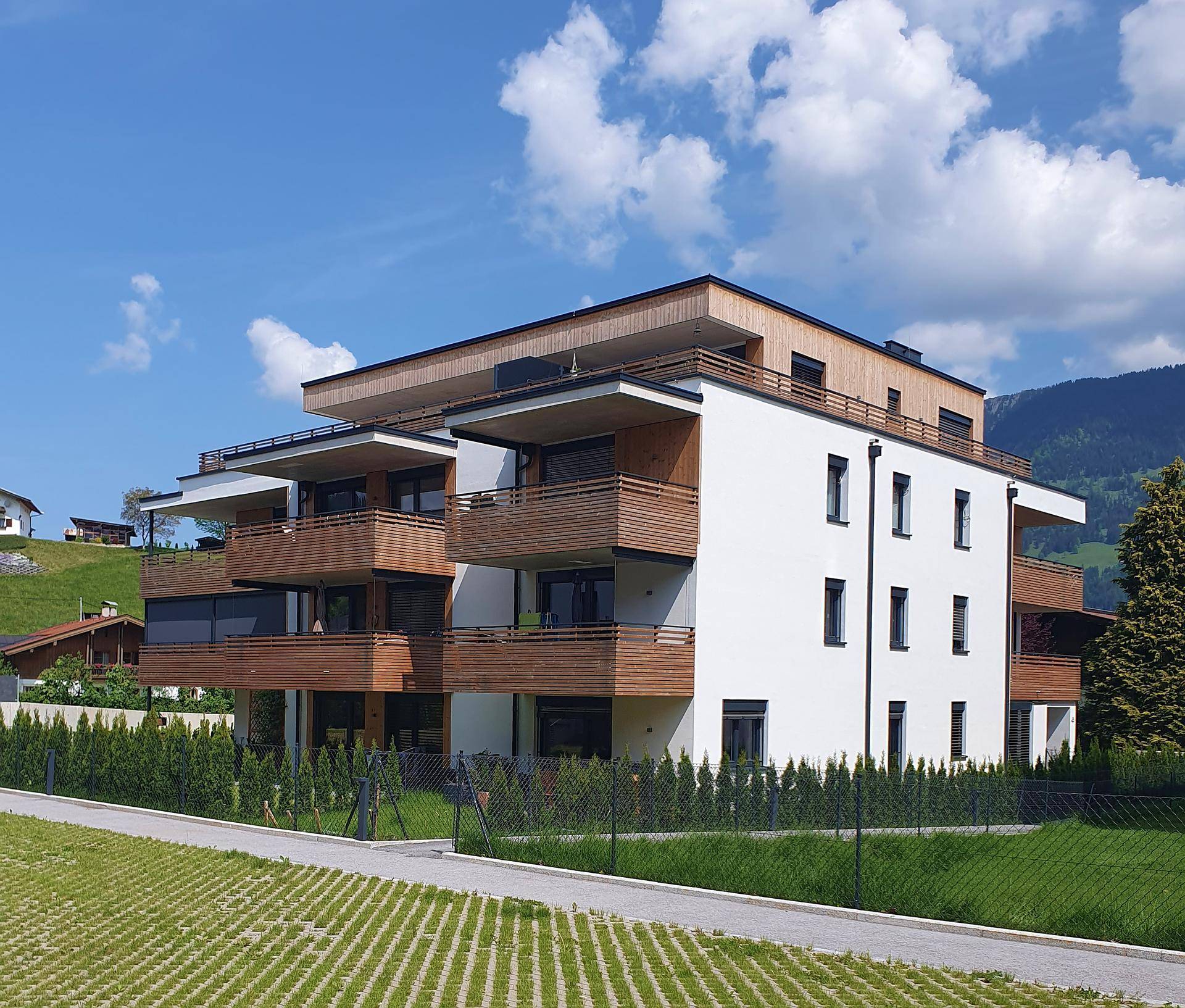 Modernes Mehrfamilienhaus mit Holzfassade, Balkonen und grüner Umgebung vor Bergkulisse.