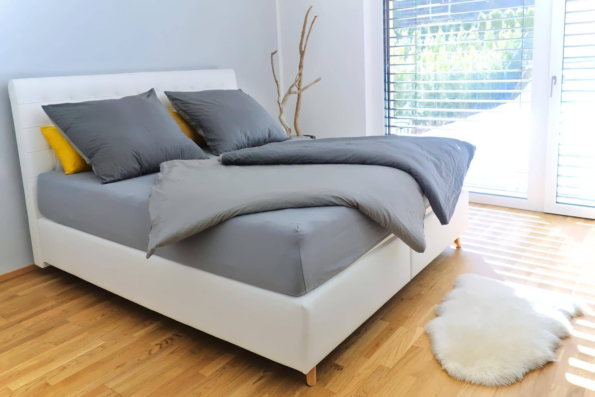 Helles Schlafzimmer mit modernem Doppelbett, Holzboden und großem Fenster mit Jalousien.