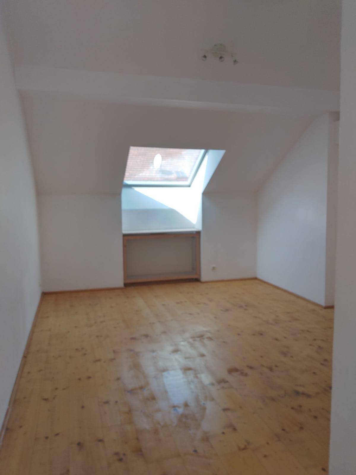 Helles Schlafzimmer mit Holzboden, weißen Wänden und einem Dachfenster, das viel Tageslicht hereinlässt.