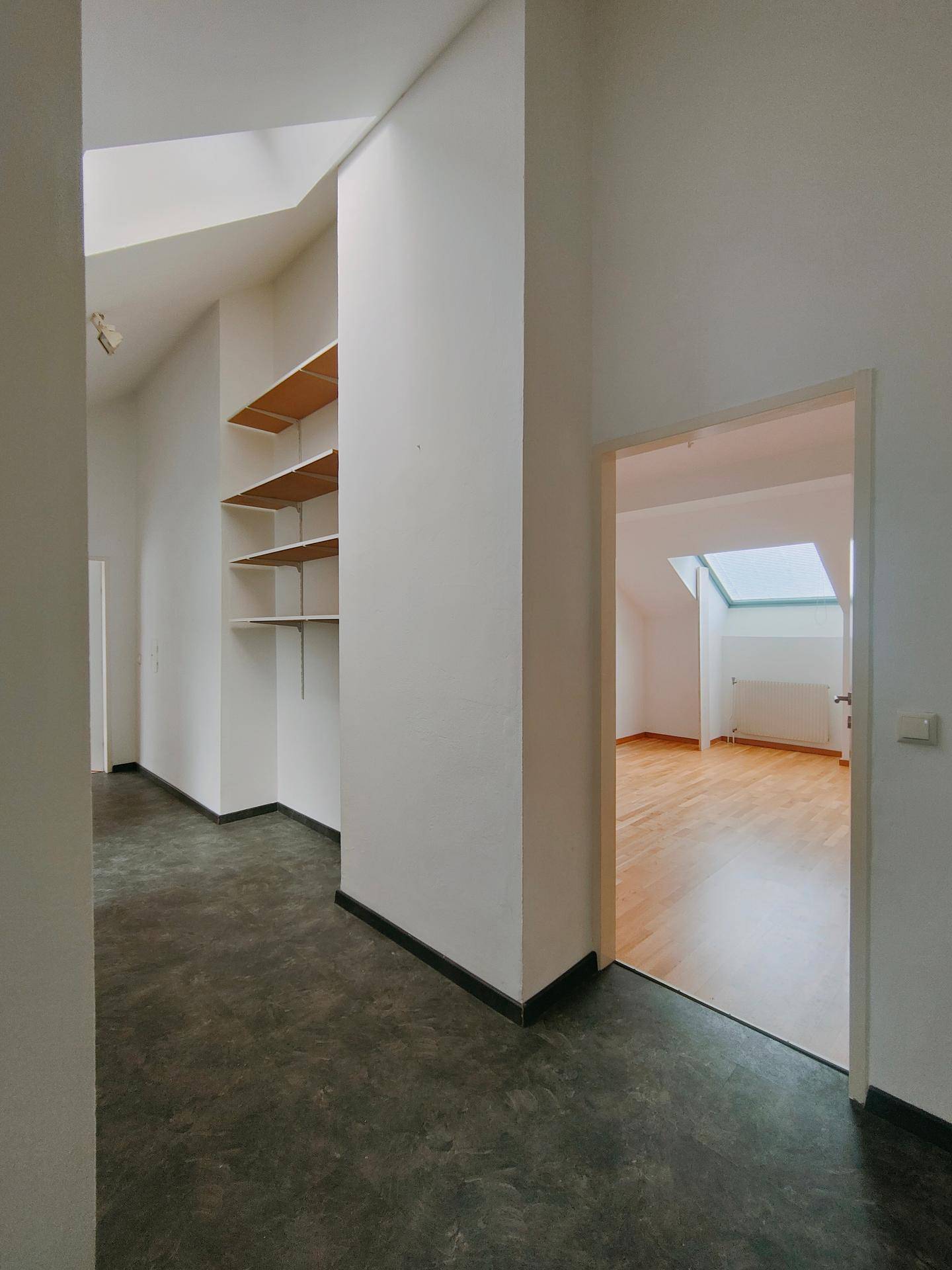 Heller Flur mit dunklem Boden, Einbauregalen und Zugang zu einem weiteren Zimmer mit Holzboden und Dachfenster.