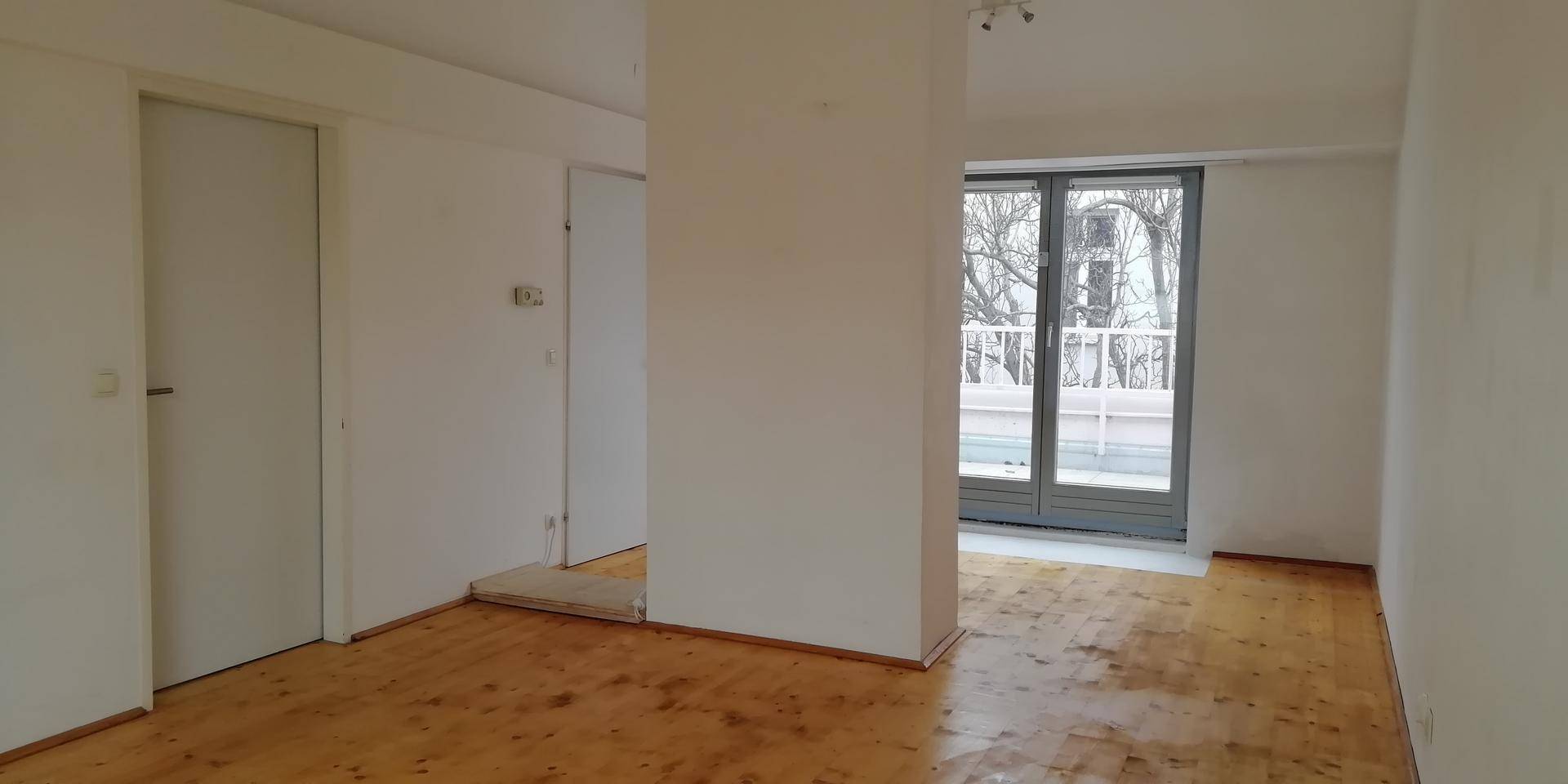 Heller, unmöblierter Raum mit Holzboden und großer Glastür, die zu einem Außenbereich führt.