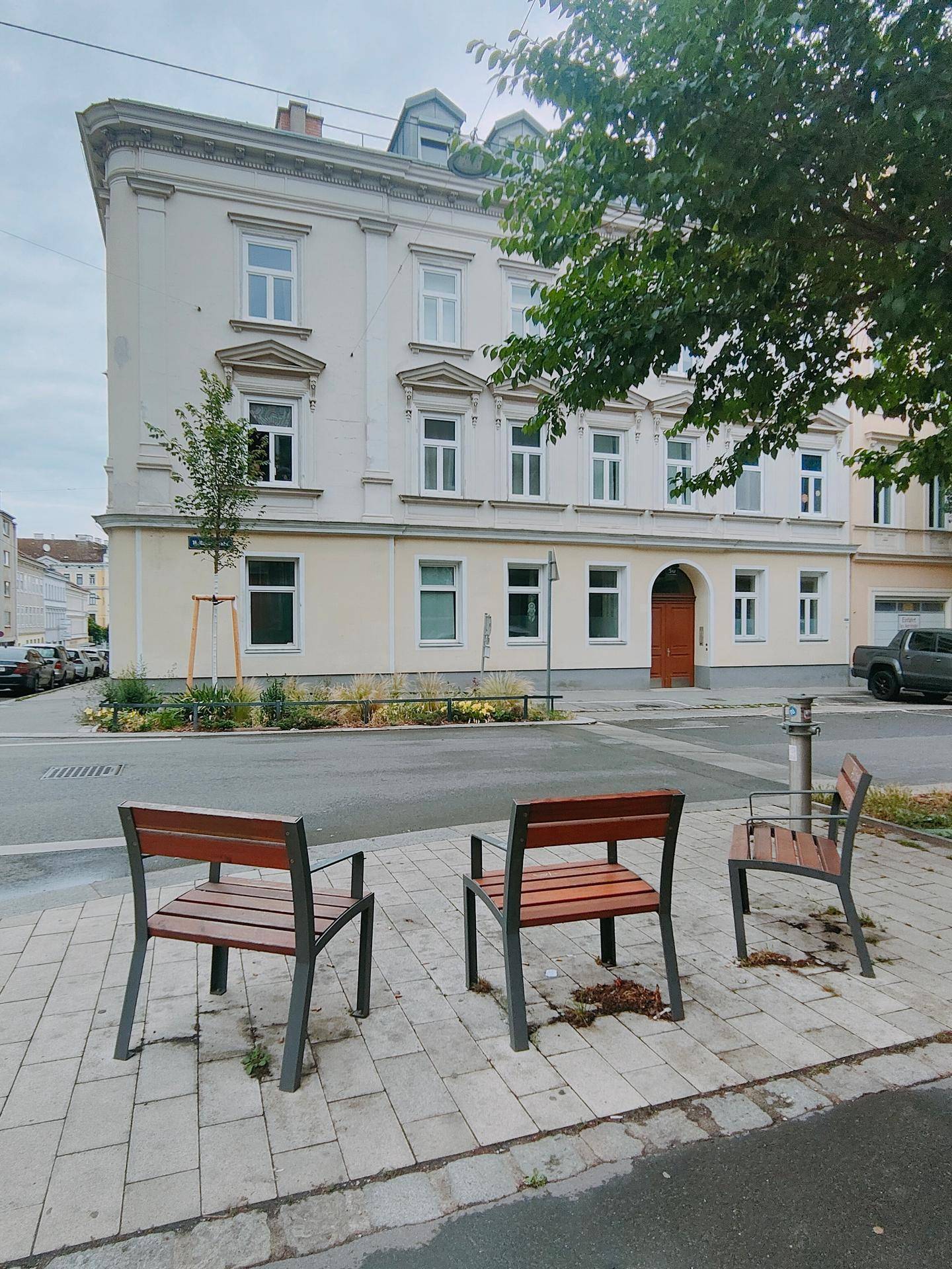 Historische Gebäudefassade an einer Straße mit Sitzbänken im Vordergrund und Bäumen.