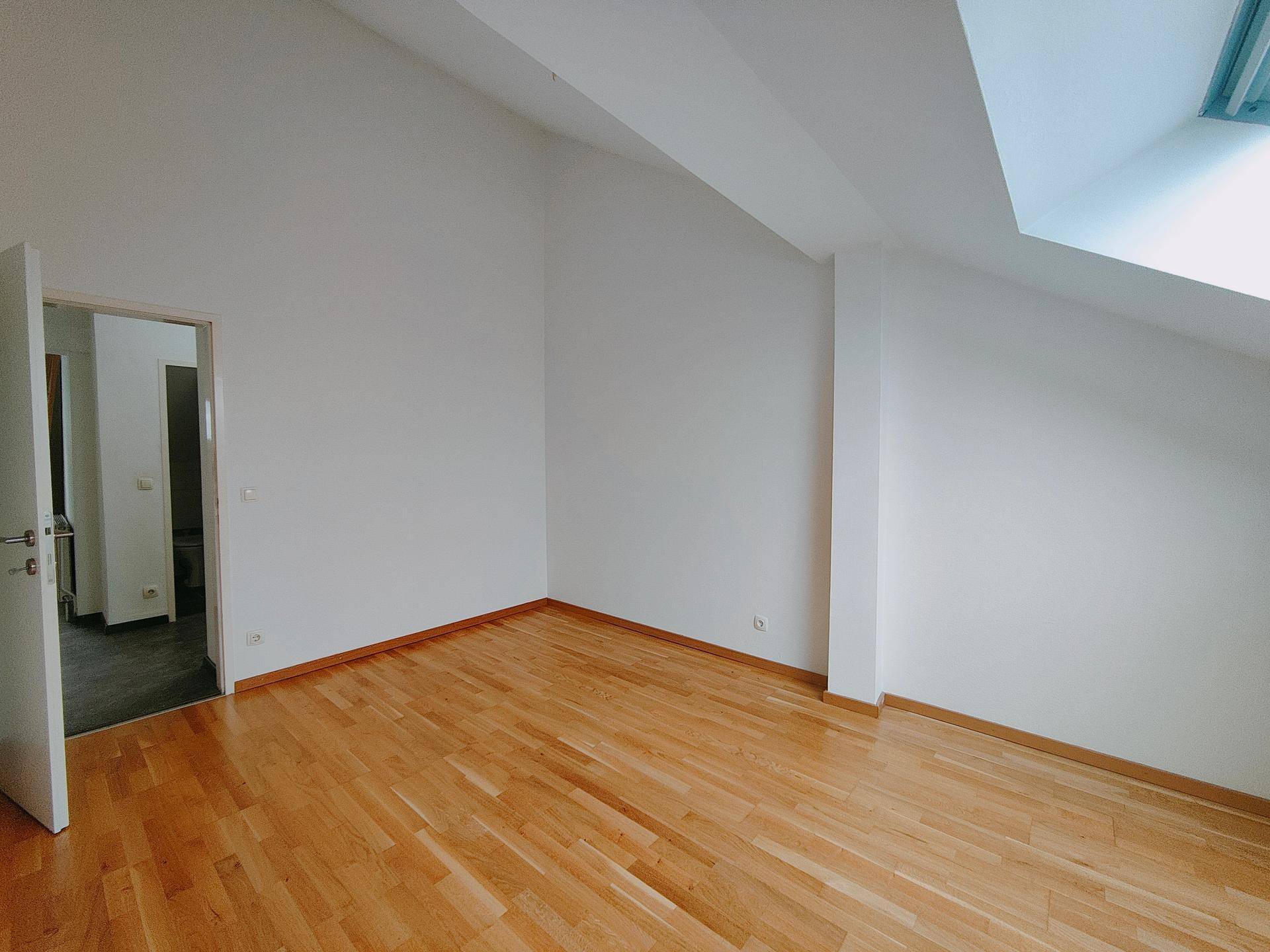 Geräumiges, unmöbliertes Zimmer mit Holzboden, weißen Wänden und einem Dachfenster, das viel Licht spendet.