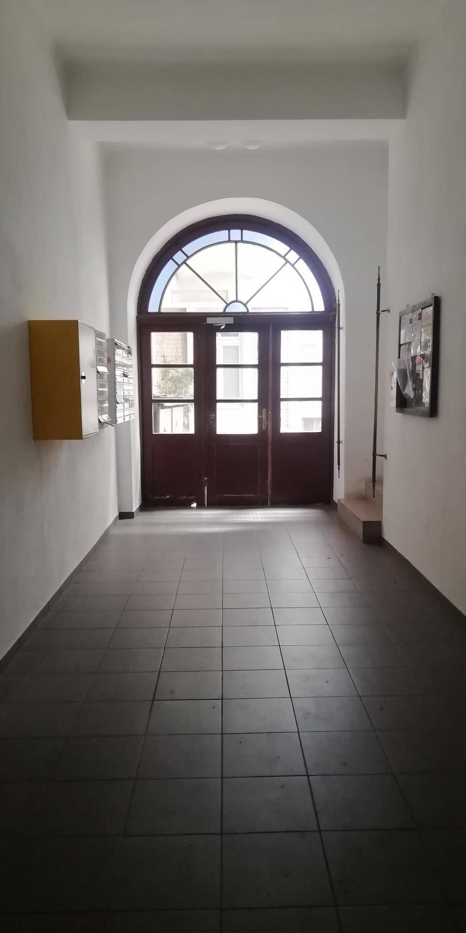Langer Eingangsbereich mit Fliesenboden und einer großen, gewölbten Holztür, die zum Gebäude führt.