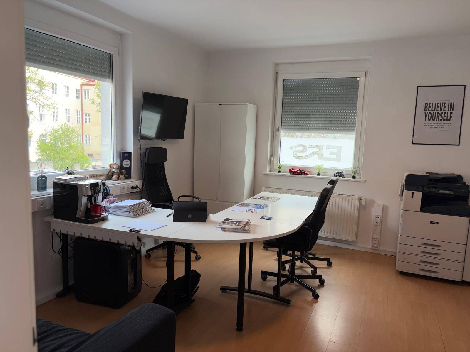 Modernes Büro mit geschwungenem Schreibtisch, Drucker und Fenstern mit Jalousien, bietet viel Arbeitsfläche und Komfort.