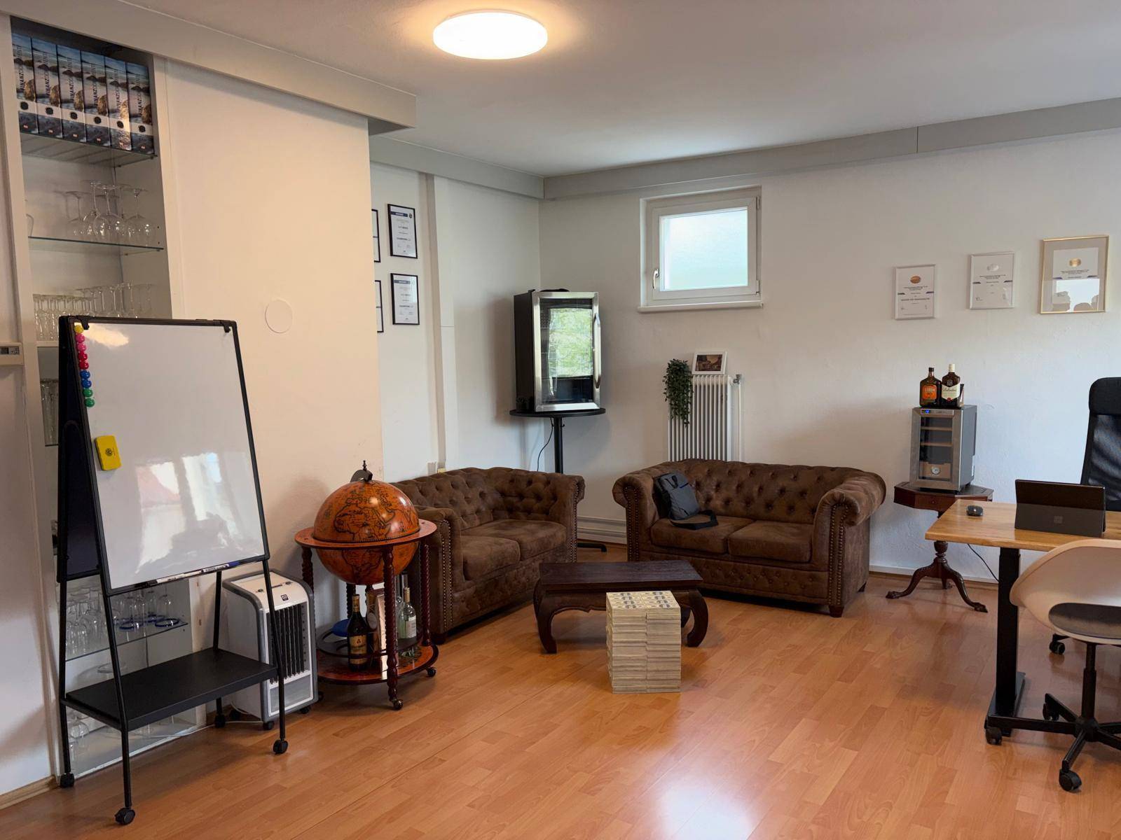 Vielseitiges Büro mit Whiteboard, bequemen Sofas und einem kleinen Fenster, geeignet für Besprechungen und Pausen.
