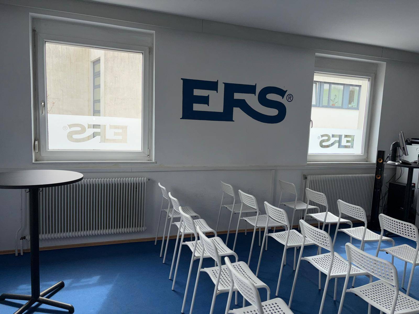 Heller Konferenzraum mit blauer Teppichboden, vielen Stühlen und einem Stehtisch, bereit für Veranstaltungen und Seminare.