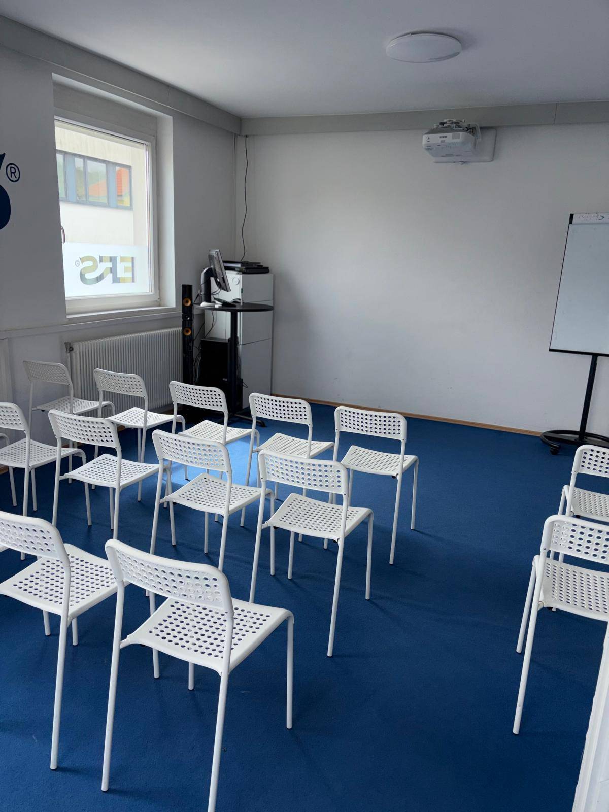 Funktionaler Konferenzraum mit blauer Teppichboden, Stuhlreihen und einem Whiteboard, ideal für Schulungen und Workshops.
