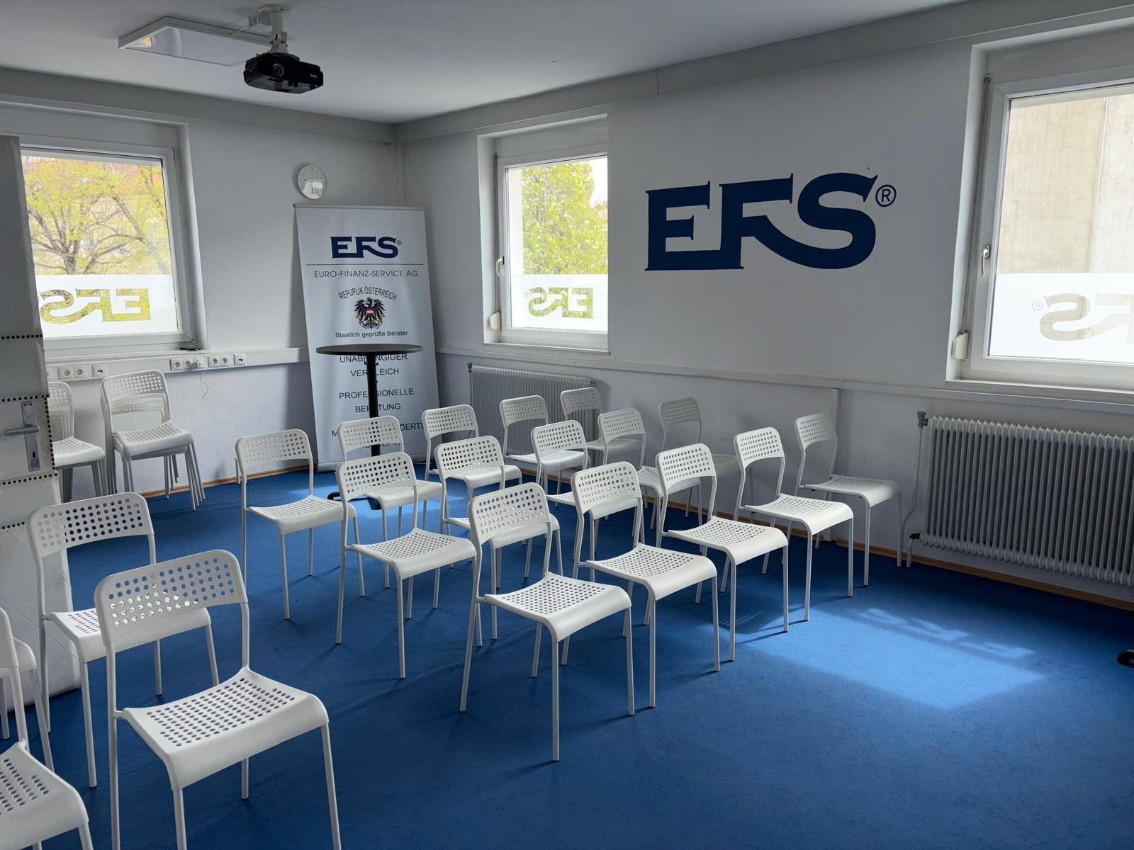 Konferenzraum mit blauer Teppichboden, Reihen von Stühlen und einem Beamer, ideal für Präsentationen und Schulungen.